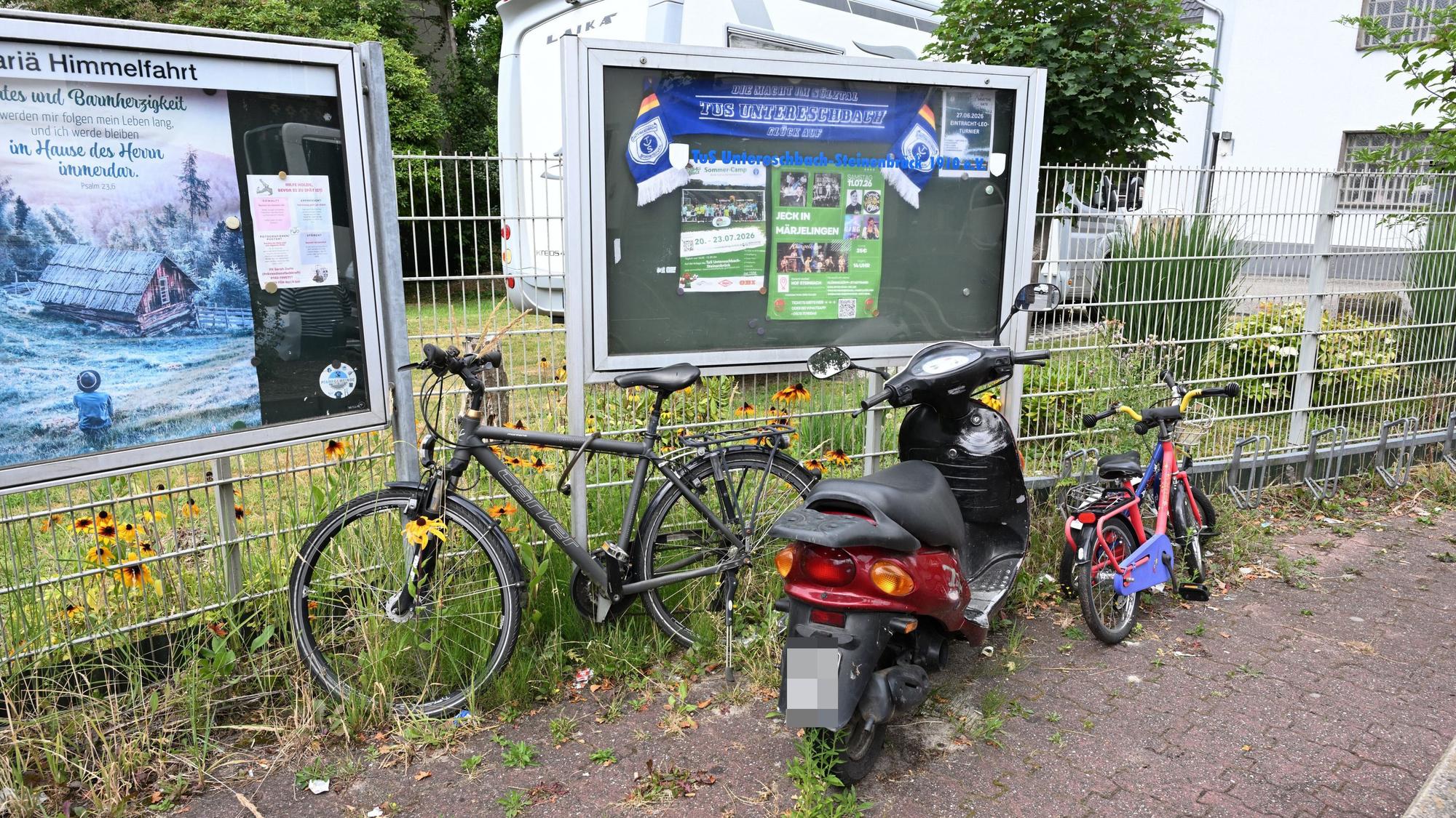 Zu sehen ist ein Fahrrad, zwei Kinderräder und ein Roller, die vor einem Zaun und zwei Infotafeln stehen.