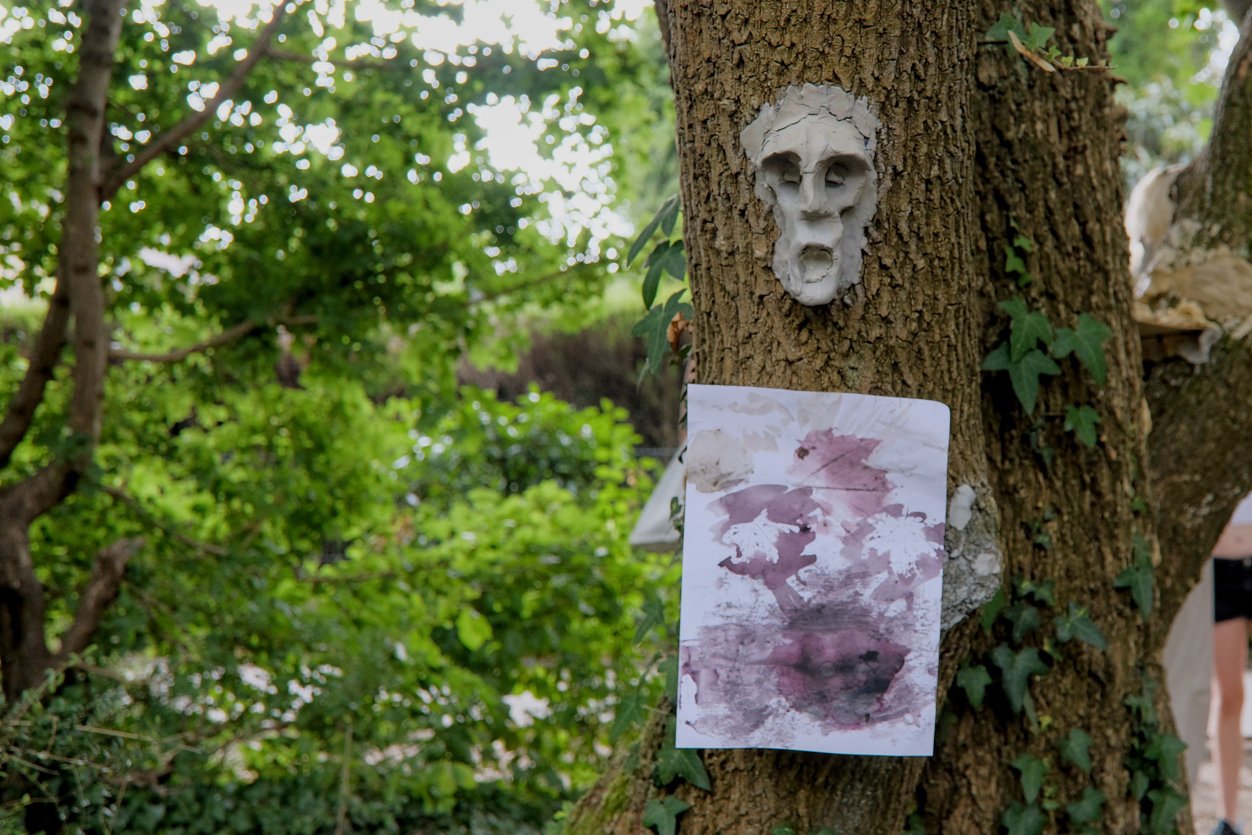 An einem Baumstamm hängt ein Poster mit Farbkleksen udn darüber eine menschliche Maske aus Ton.
