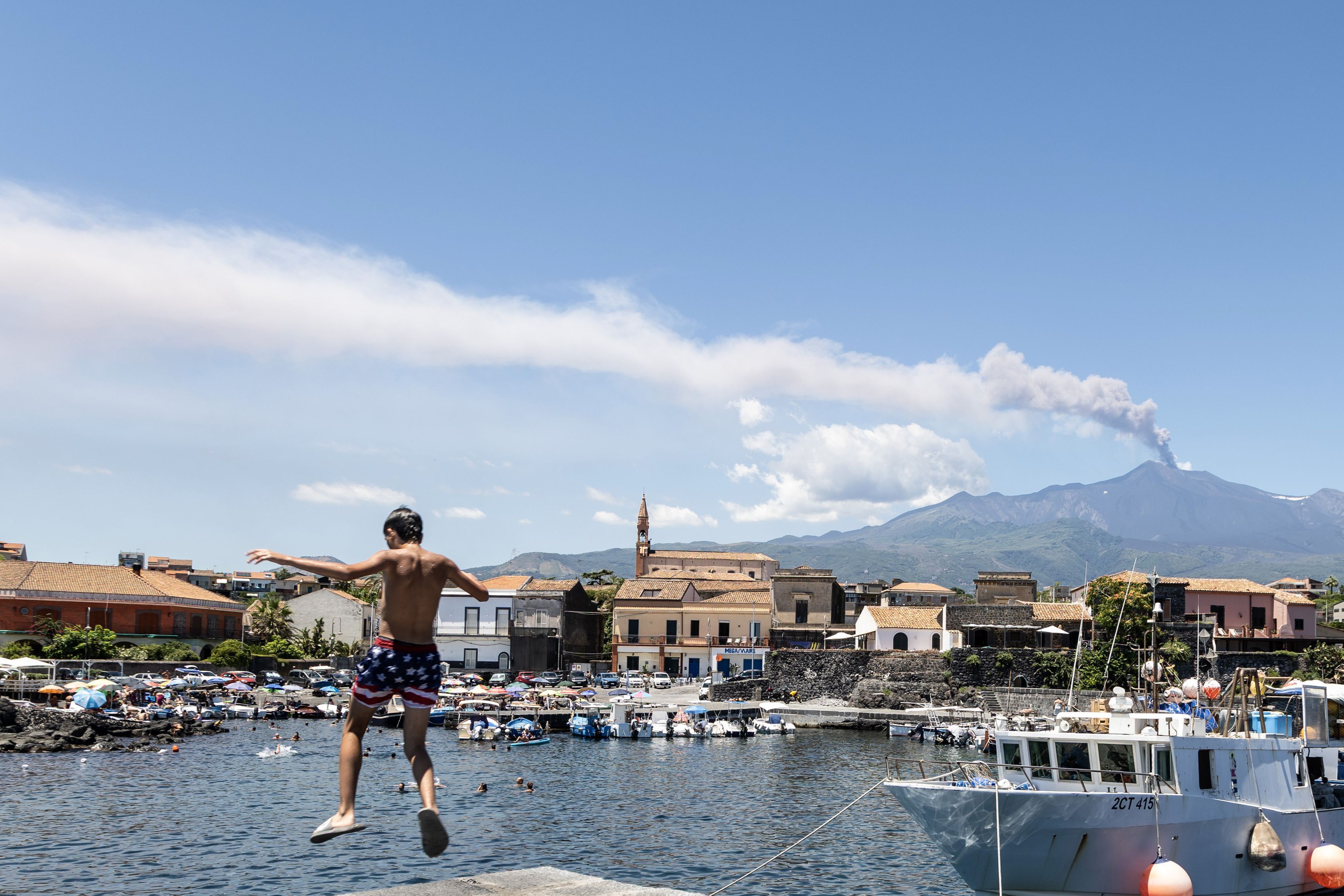Blick auf eine Aschewolke des Vulkans Ätna, während ein Kind am Hafen ins Wasser springt. Wegen des starken Ascheausstoßes des Ätna auf der italienischen Mittelmeerinsel Sizilien ist der Flugverkehr am Flughafen Catania derzeit nur bedingt möglich.