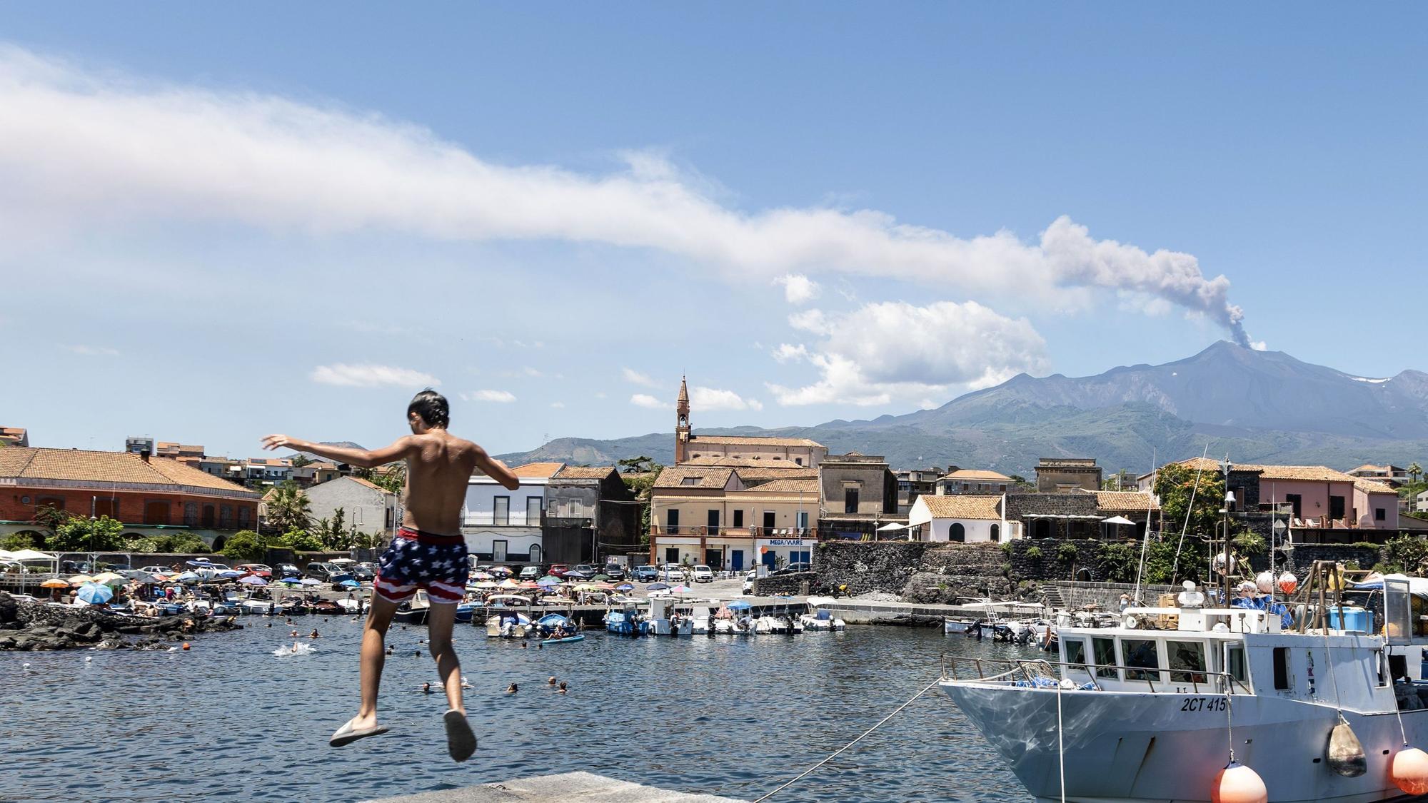 Blick auf eine Aschewolke des Vulkans Ätna, während ein Kind am Hafen ins Wasser springt. Wegen des starken Ascheausstoßes des Ätna auf der italienischen Mittelmeerinsel Sizilien ist der Flugverkehr am Flughafen Catania derzeit nur bedingt möglich.