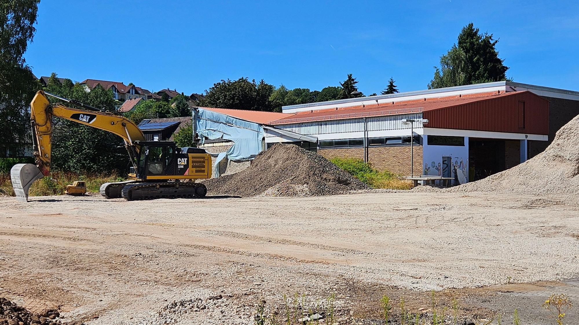 Ein Bagger steht auf einer befestigten Fläche. Dahinter ist die Turnhalle zu sehen.