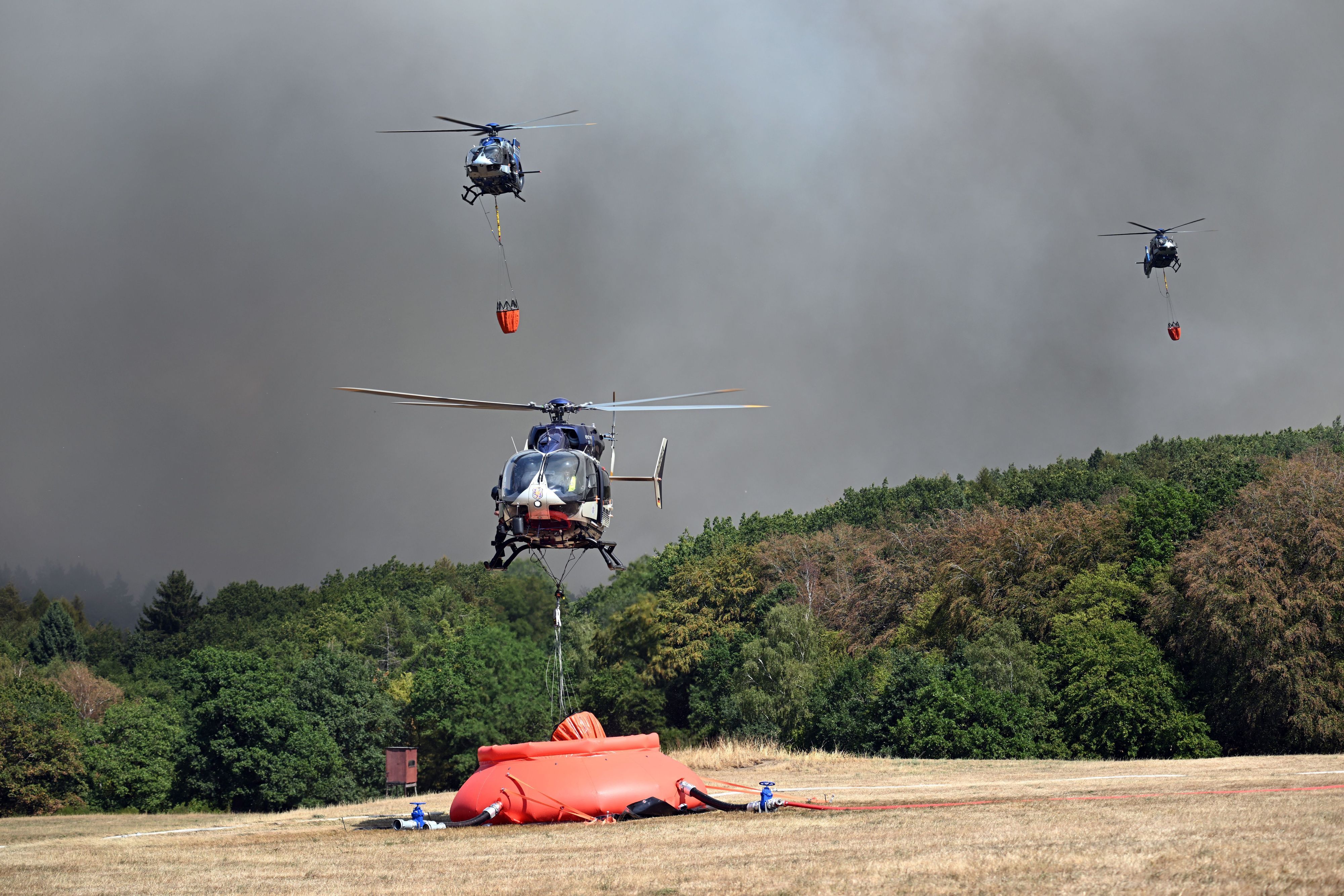 Ein Polizeihubschrauber mit Löschwasser-Außenlastbehälter startet zum Einsatz bei den Löscharbeiten beim Waldbrand im Hürtgenwald. Weitere Hubschrauber ssind im Anflug.