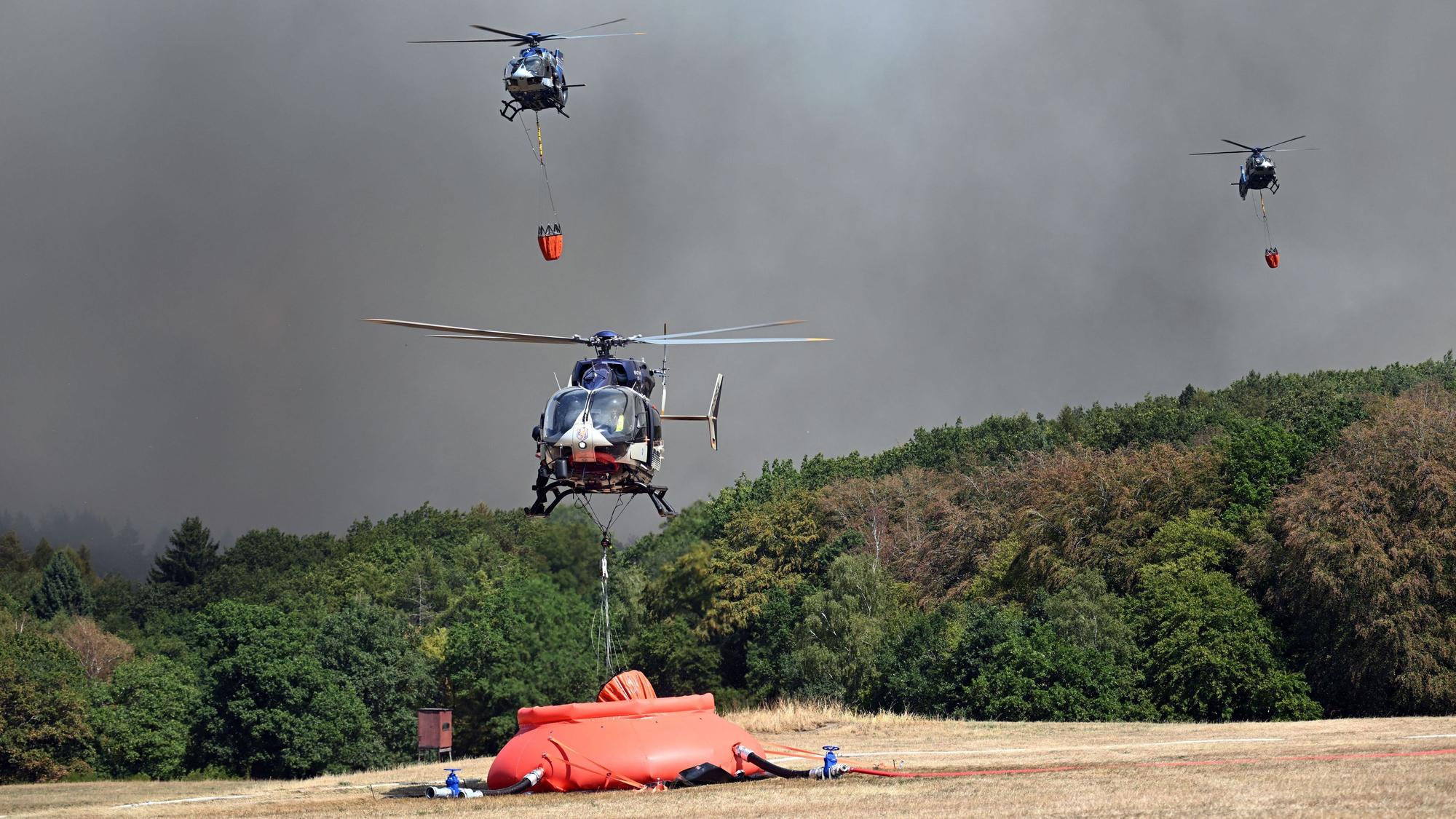 Ein Polizeihubschrauber mit Löschwasser-Außenlastbehälter startet zum Einsatz bei den Löscharbeiten beim Waldbrand im Hürtgenwald. Weitere Hubschrauber ssind im Anflug.
