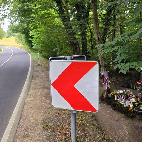Eine Warntafel, eine Kreuz und Blumen stehen neben der Landstraße im Dürschtal bei Kürten-Dürscheid, an der eine Leitplanke fehlt.