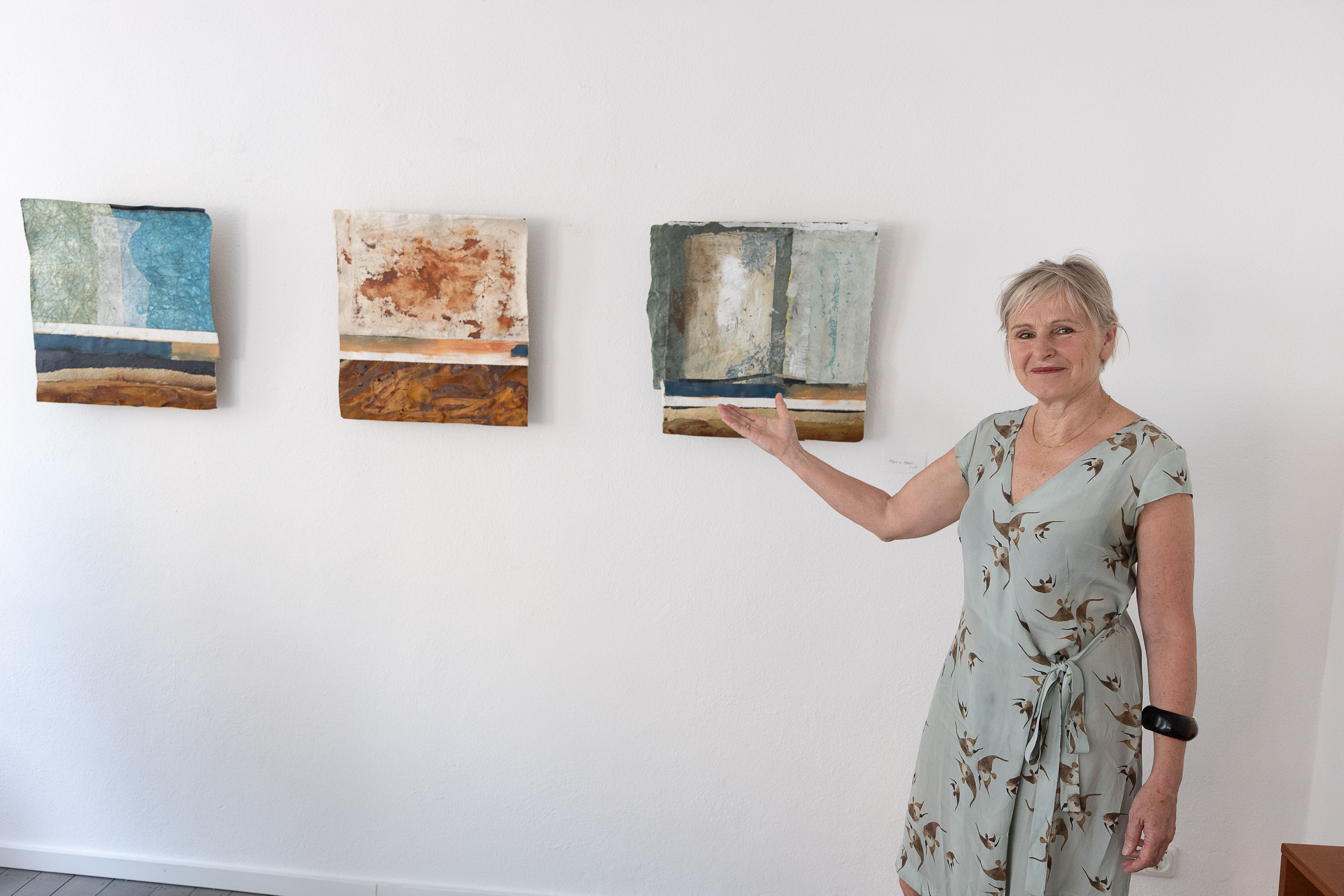 Die Künstlerin Heike Hofmann steht vor einer Wand, an der drei ihrer Bilder hängen. Sie zeigt mit der rechten Hand darauf.
