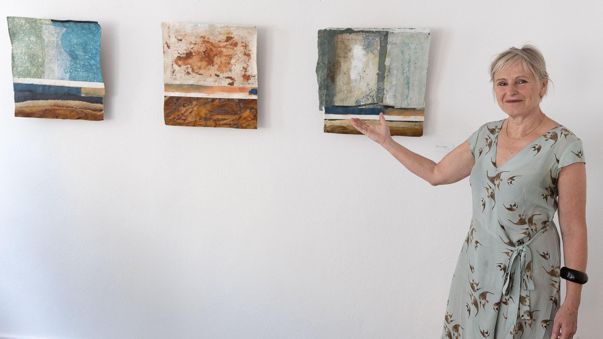 Die Künstlerin Heike Hofmann steht vor einer Wand, an der drei ihrer Bilder hängen. Sie zeigt mit der rechten Hand darauf.