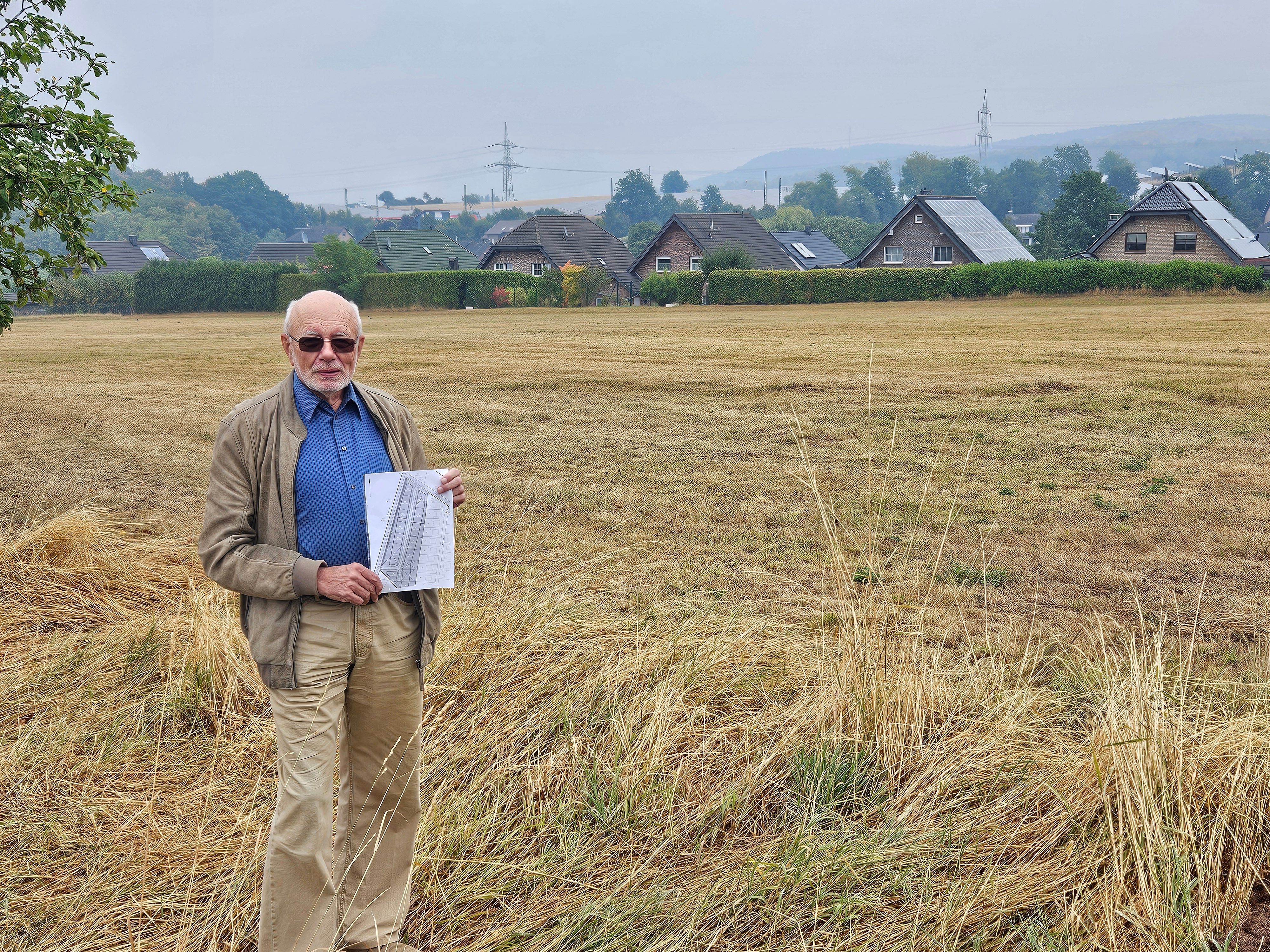Hans Reiff steht mit einem Plan vor einer Wiese. Im Hintergrund sind Häuser zu sehen.