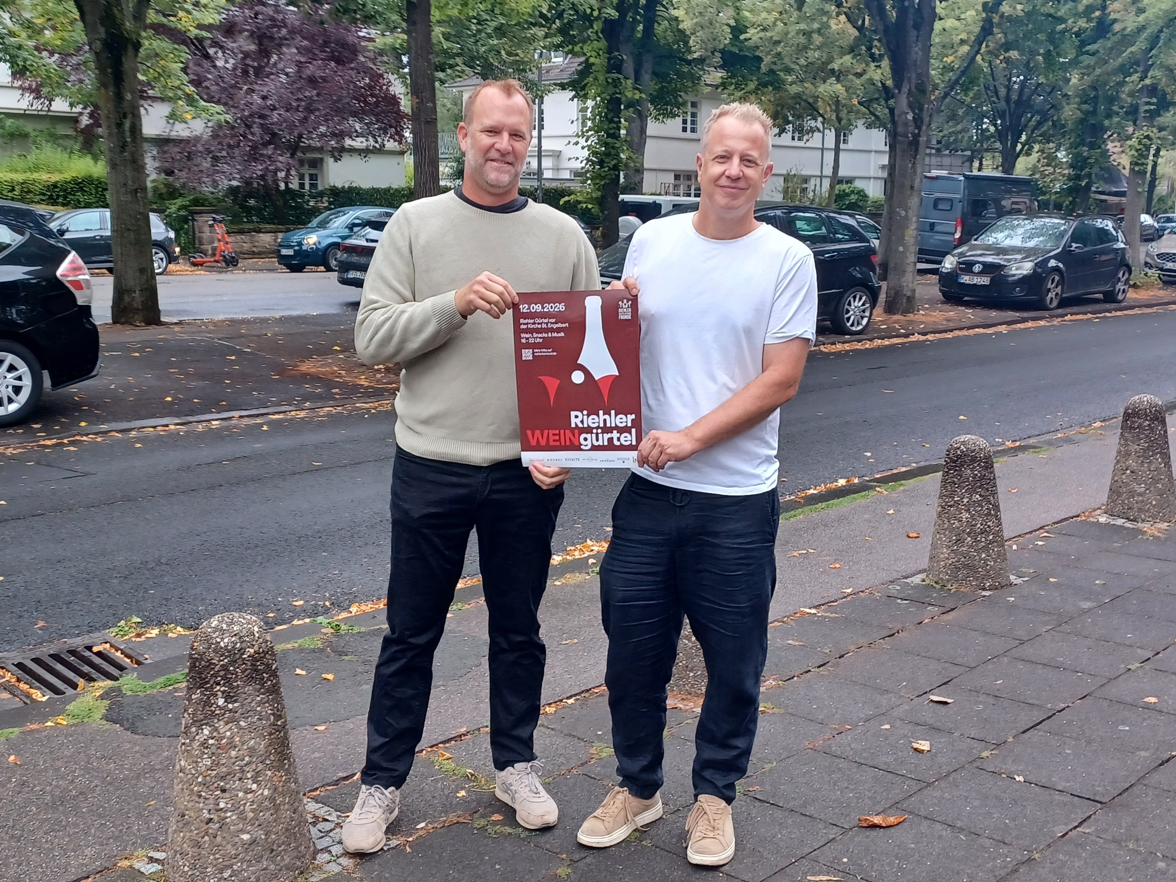 Zwei Männer stehen auf einem Bürgersteig und halten ein Plakat in den Händen, auf dem steht: Riehler WEINgürtel.