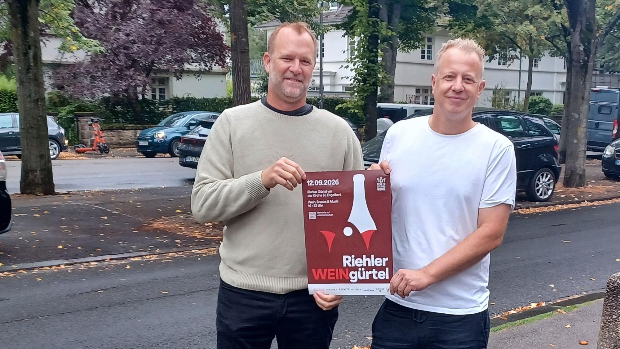 Zwei Männer stehen auf einem Bürgersteig und halten ein Plakat in den Händen, auf dem steht: Riehler WEINgürtel.