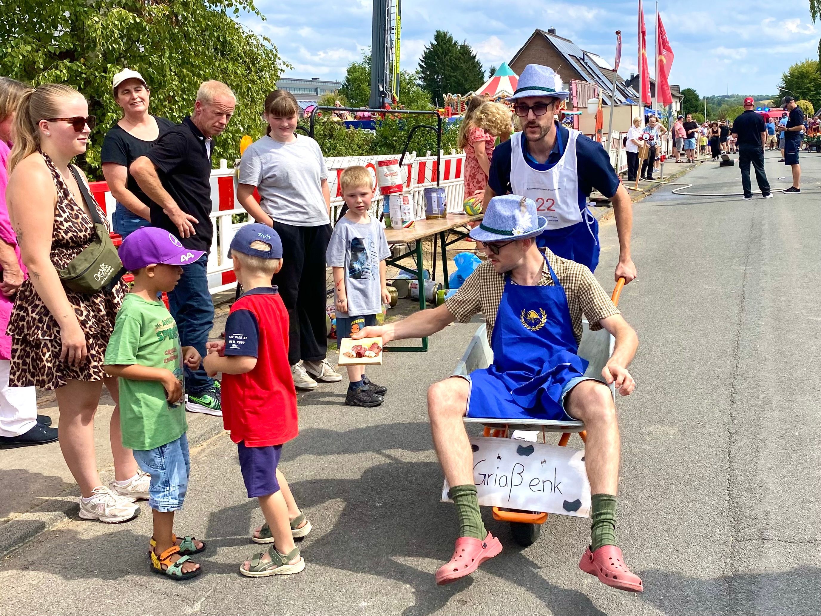 Ein Mann mit lustigem Hut sitzt in einer Schubkarre, die ein zweiter Mann schiebt. Am Straßenrand Kinder, denen einer der Männer eine Schokolade hinhält.