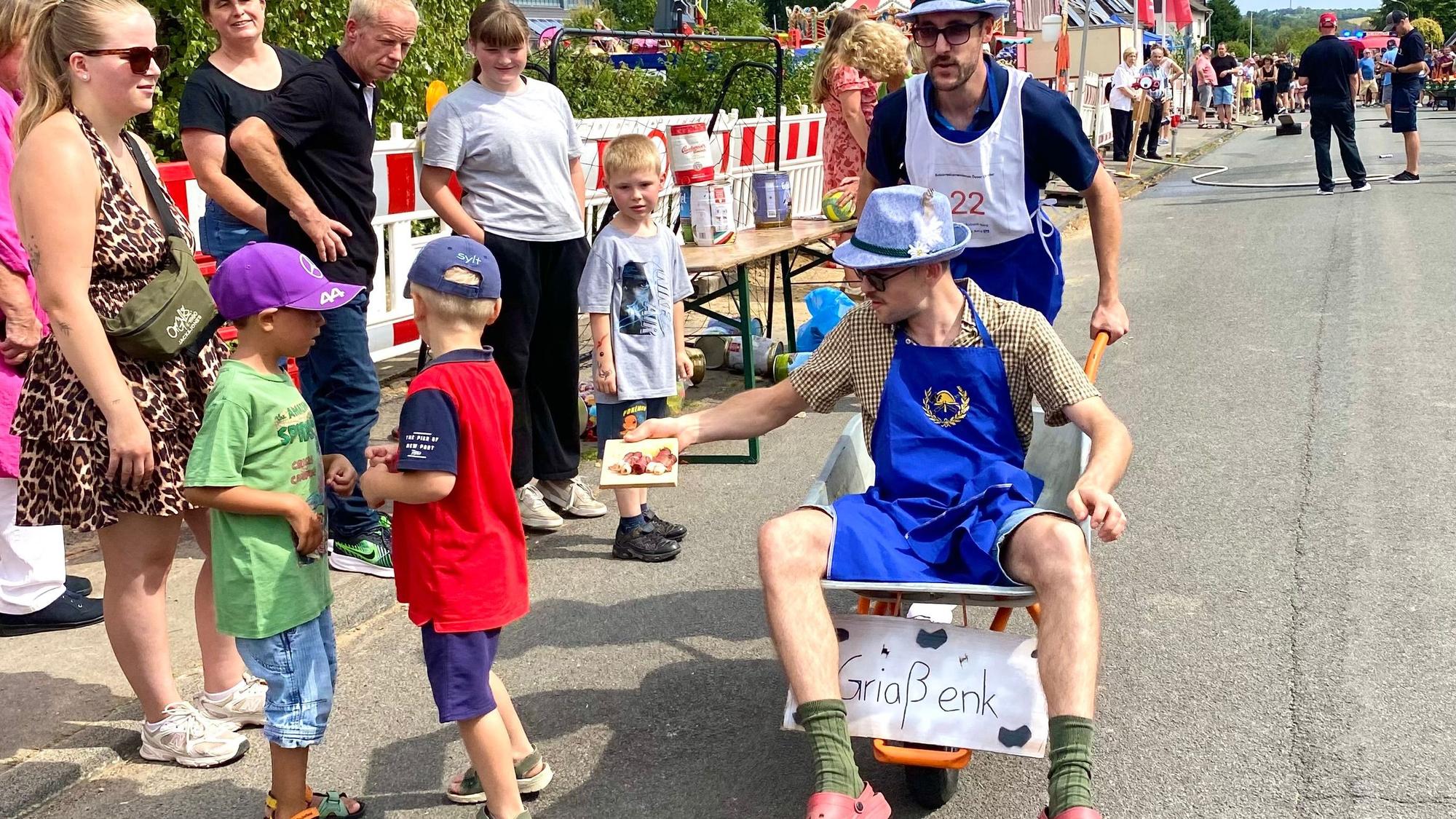 Ein Mann mit lustigem Hut sitzt in einer Schubkarre, die ein zweiter Mann schiebt. Am Straßenrand Kinder, denen einer der Männer eine Schokolade hinhält.