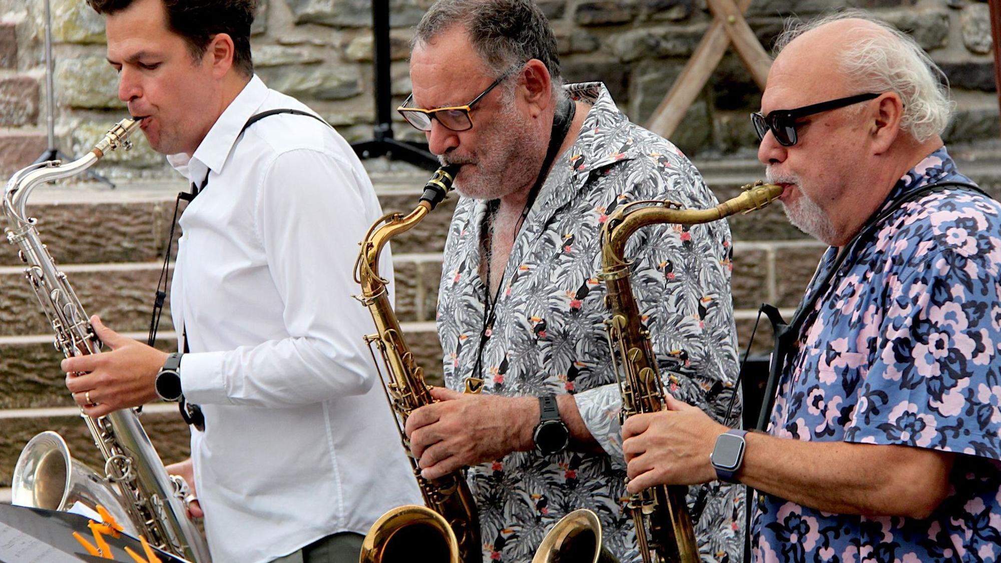 Drei Männer, zwei davon mit Brillen, stehen nebeneinander und spielen Saxofon.