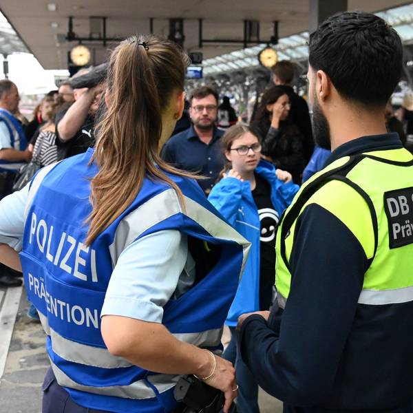 Zwei Sicherheitsbeamte der Deutschen Bahn und der Polizei stehen an einem vollen Bahngleis mit dem Rücken zur Kamera.