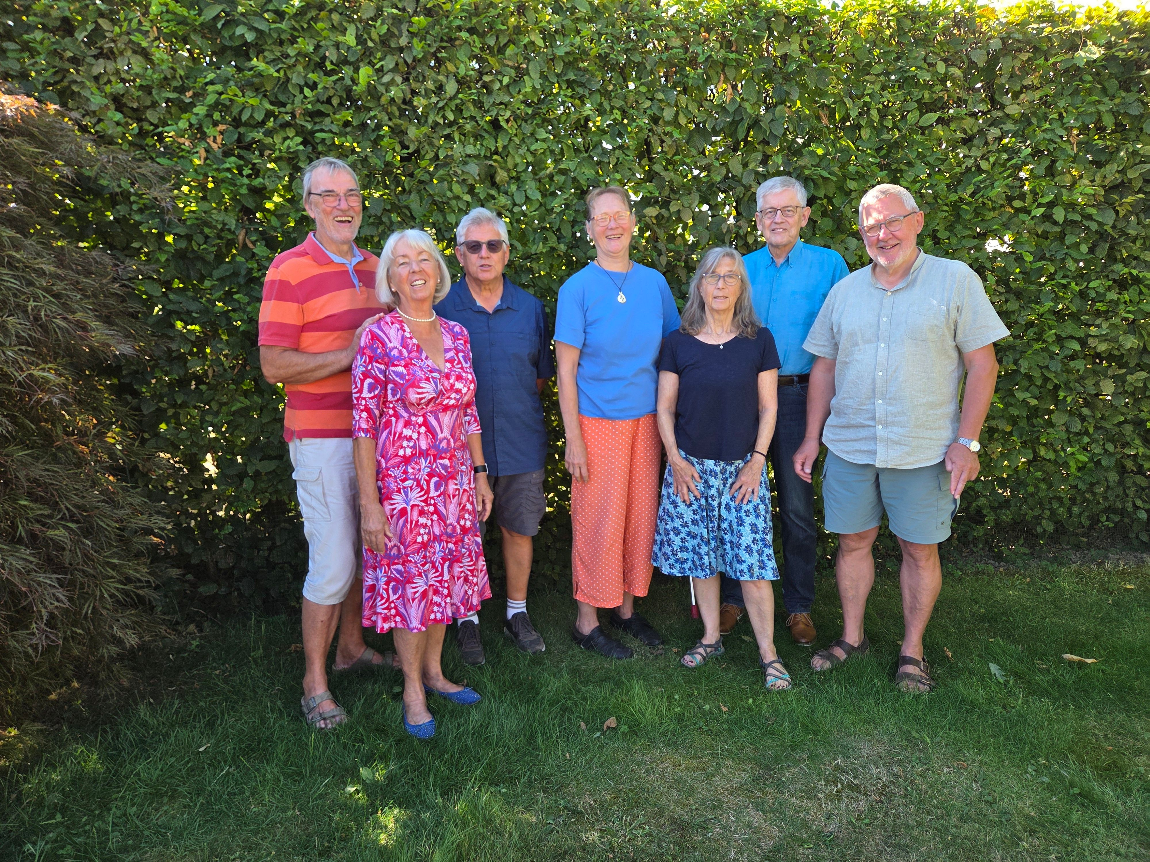 Eine Gruppe gut gelaunter Menschen steht vor einer Hecke auf einem Rasen und lächelt.