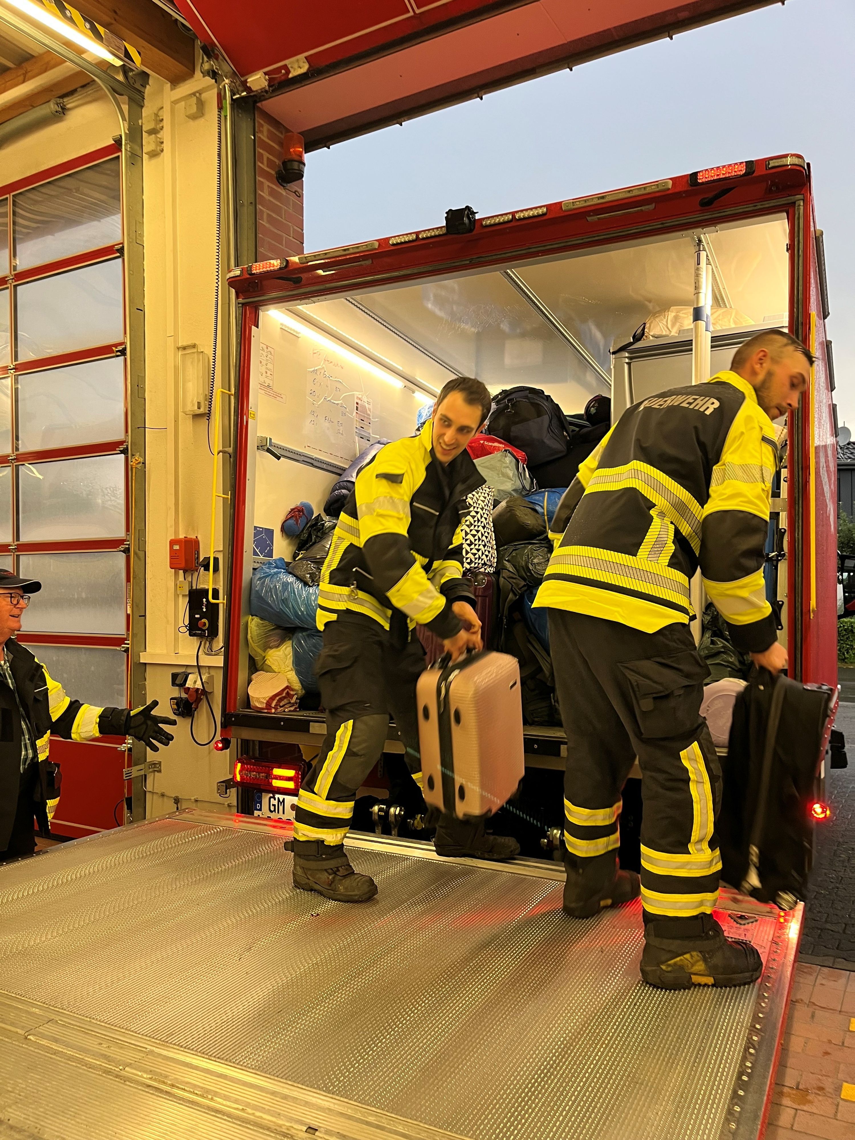 Zwei Feuerwehrmänner laden Reisegepäck aus einem Feuerwehrfahrzeug aus.