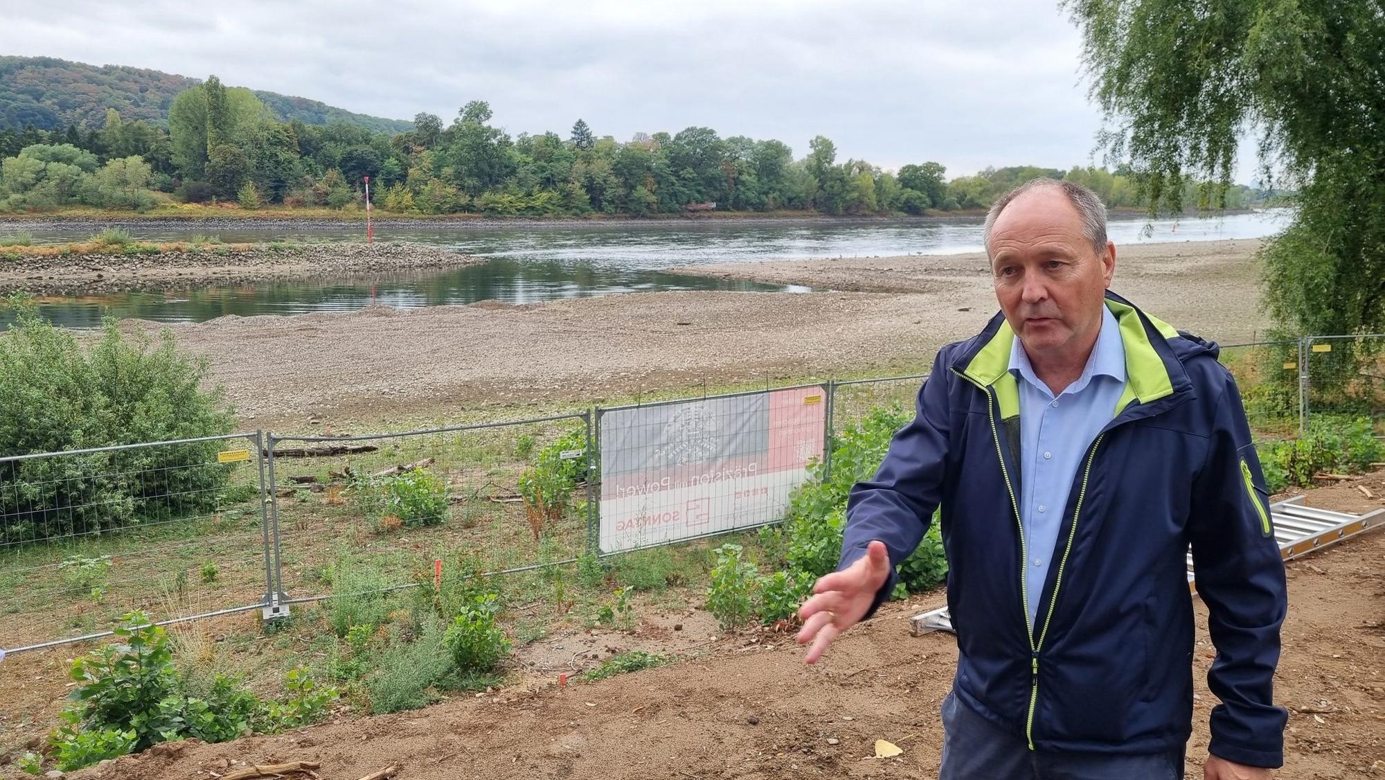 Ein Mann mit blauer Jacke steht neben einem Bauzaun am Ufer des Rheins.