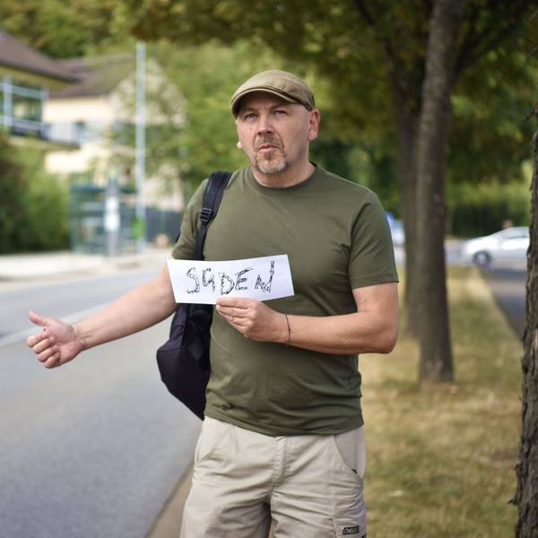 Thorsten Wirtz steht in grünem T-Shirt mit Rucksack und Schirmmütze an einer Straße und hält einen Daumen nach oben. In der anderen Hand hält er ein Schild auf dem "Süden" steht.