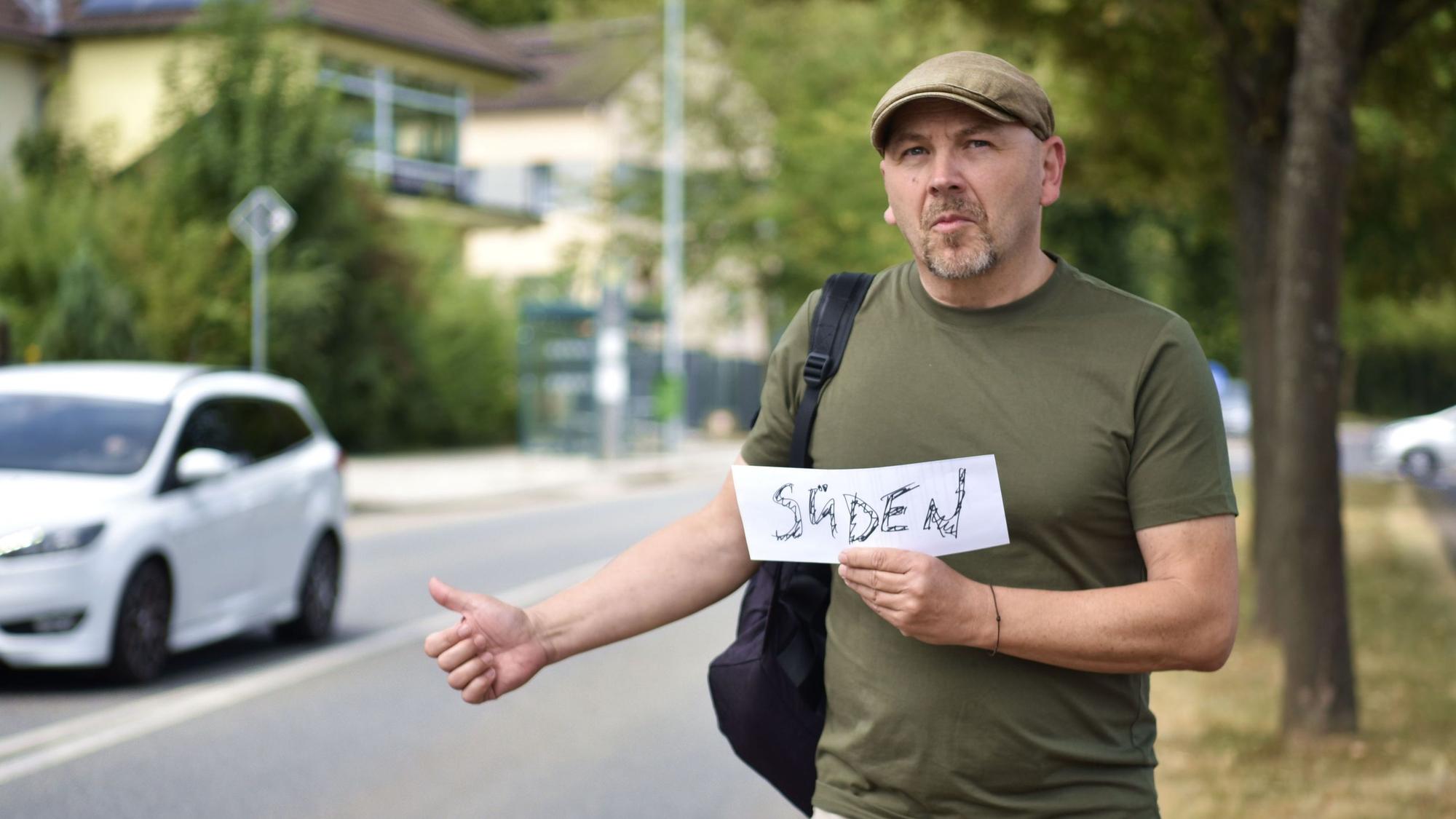 Thorsten Wirtz steht in grünem T-Shirt mit Rucksack und Schirmmütze an einer Straße und hält einen Daumen nach oben. In der anderen Hand hält er ein Schild auf dem "Süden" steht.