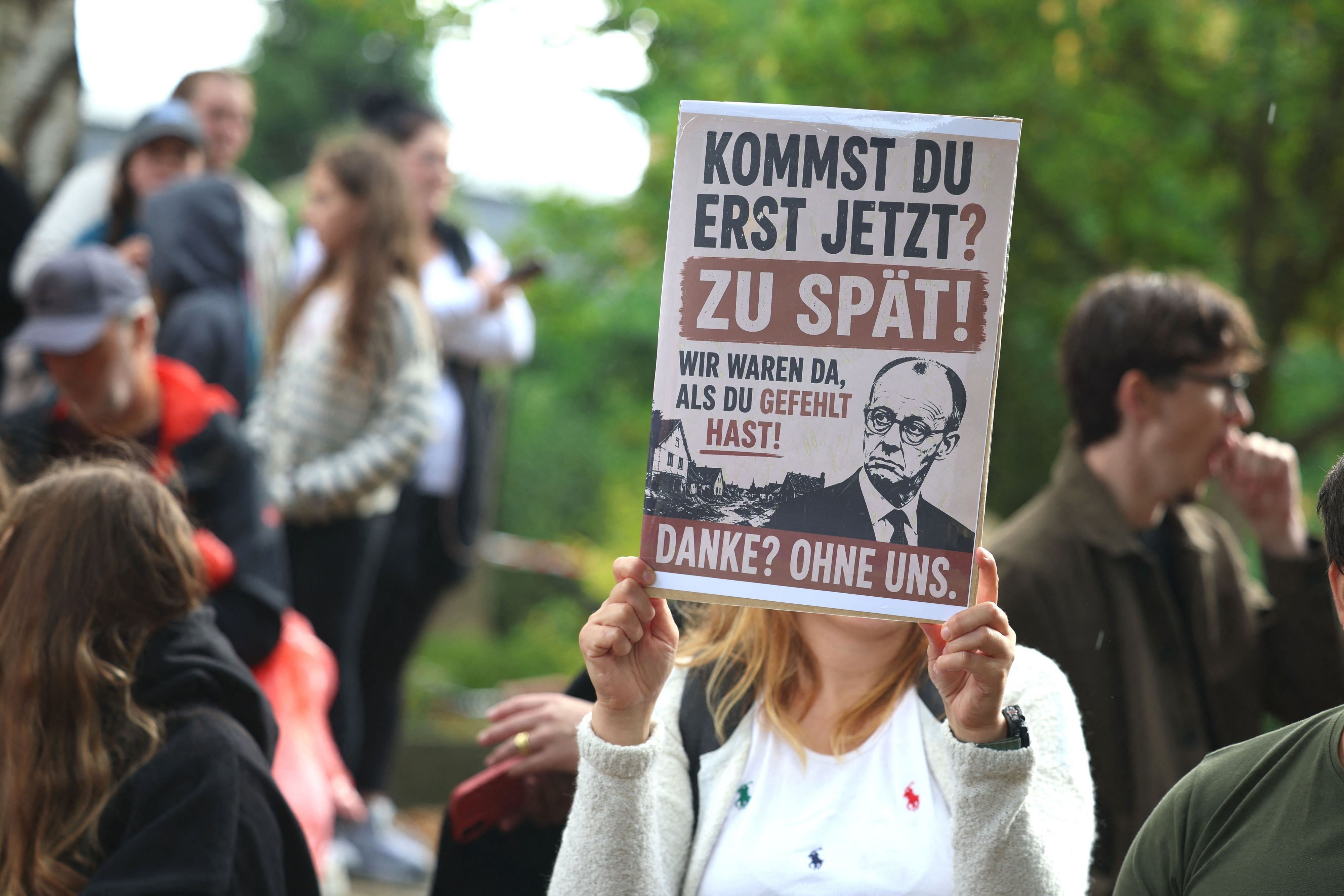 Eine Frau hält ein Plakat mit der Aufschrift „Kommst du erst jetzt? Zu spät!“ bei einem Besuch von Bundeskanzler Merz in Hürtgenwald-Gey.