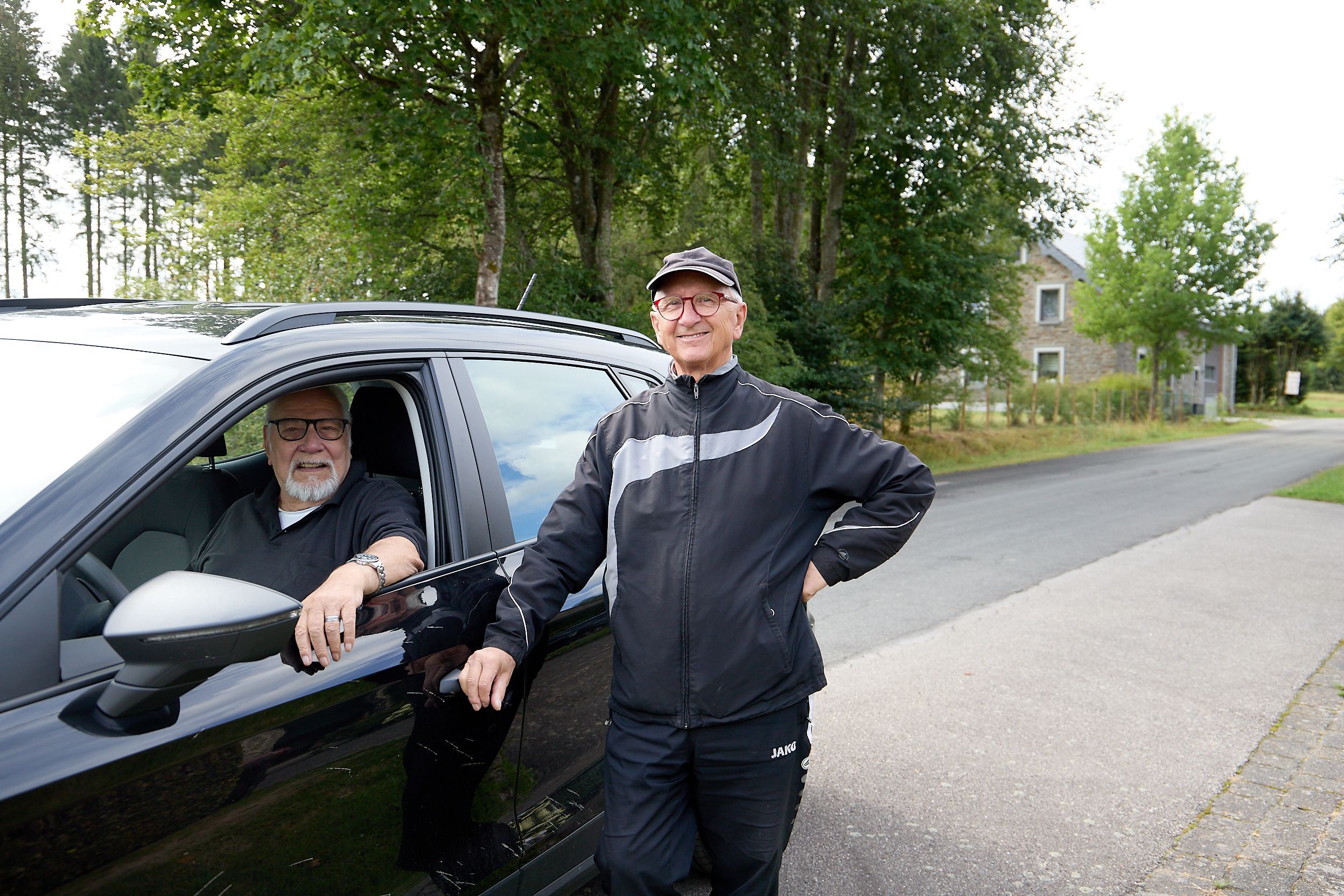 Das Foto zeigt zwei ältere Männer: Einer sitzt bei geöffnetem Fenster am Steuer eines schwarzen Autos, der andere steht daneben und lehnt sich mit einem Arm an das Fahrzeug.