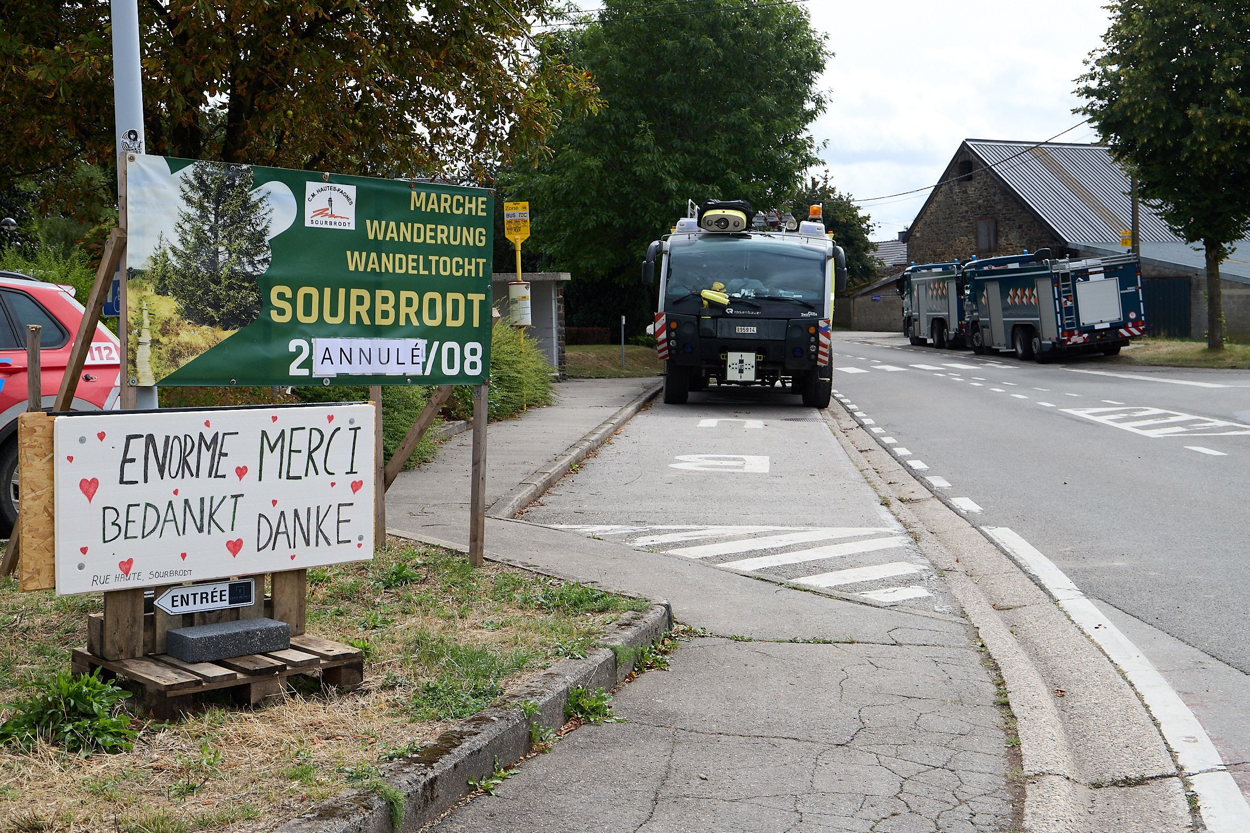 In Sourbrodt parken zwei große Einsatzfahrzeuge am Straßenrand, im Vordergrund sind Schilder zu sehen, die auf eine abgesagte Veranstaltung hinweisen und in mehreren Sprachen den Einsatzkräften gedankt wird.