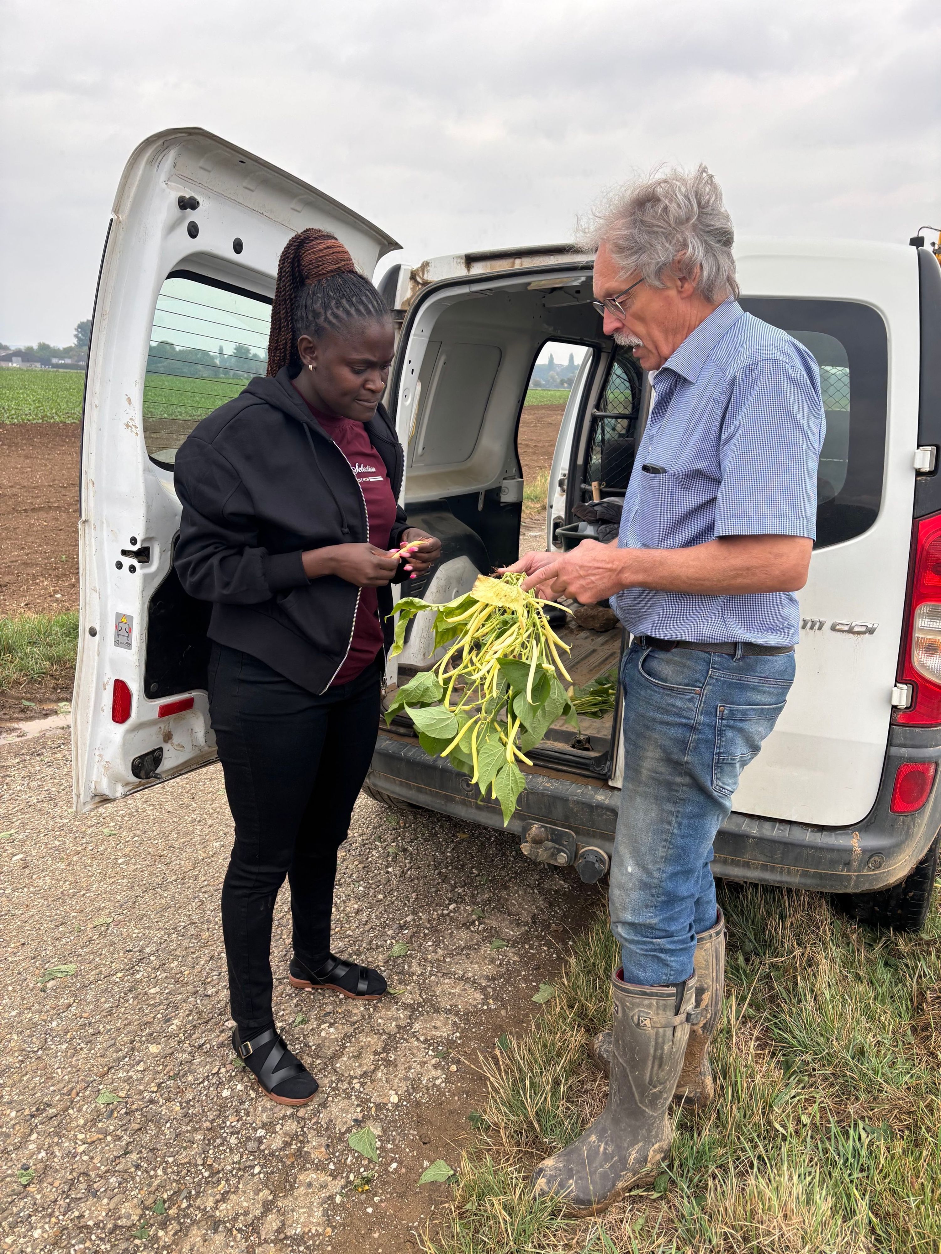 Bridgit Obuya und Hermann-Josef Weinberg stehen an einem Auto, er hält Pflanzenteile in der Hand.