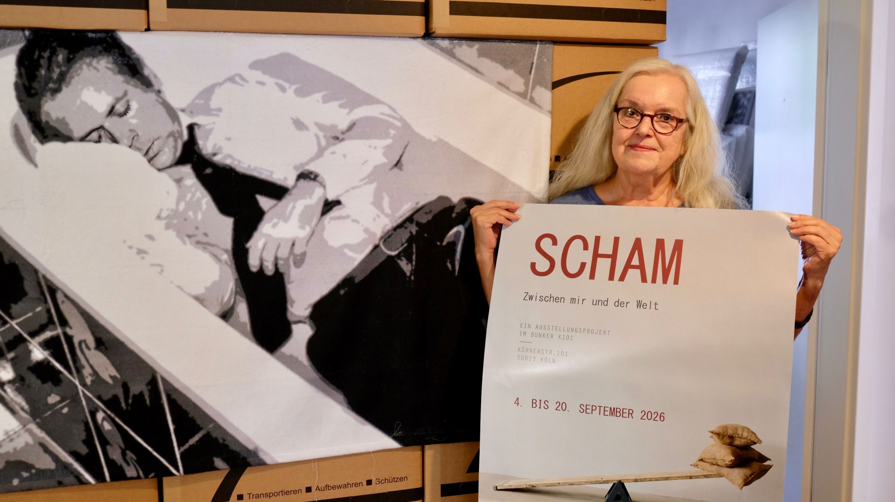 Eine Frau mit blondem Haar und Brille hält ein Veranstaltungsplakat. Hinter ihr ist ein großes Schwarz-Weiß-Bild zu sehen. Es zeigt Uwe Barschel in der Badewanne.