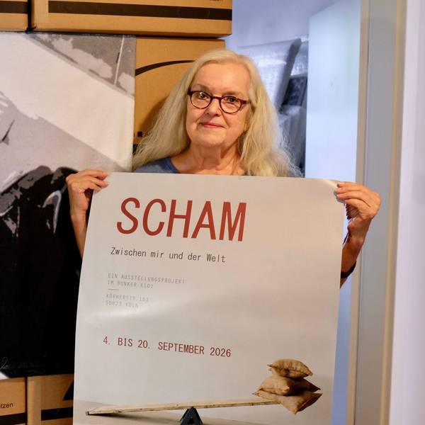 Eine Frau mit blondem Haar und Brille hält ein Veranstaltungsplakat. Hinter ihr ist ein großes Schwarz-Weiß-Bild zu sehen. Es zeigt Uwe Barschel in der Badewanne.