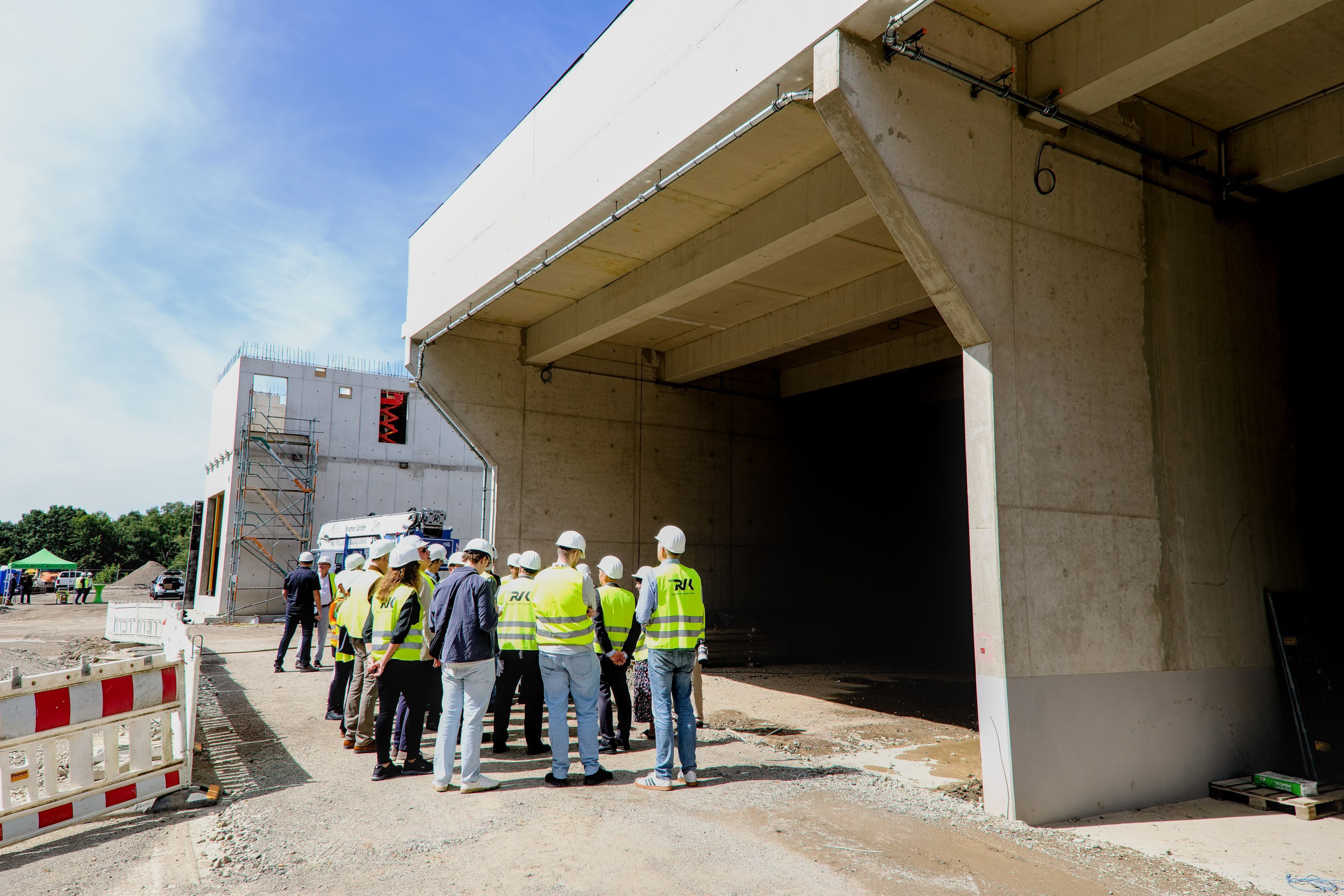 Zu sehen ist eine Gruppe Menschen, die unter dem Rohbau für ein großes Bauwerk steht.