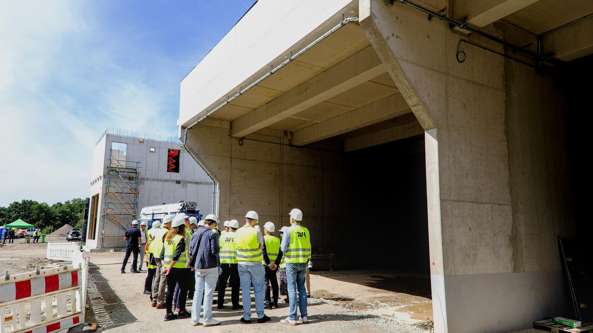 Zu sehen ist eine Gruppe Menschen, die unter dem Rohbau für ein großes Bauwerk steht.
