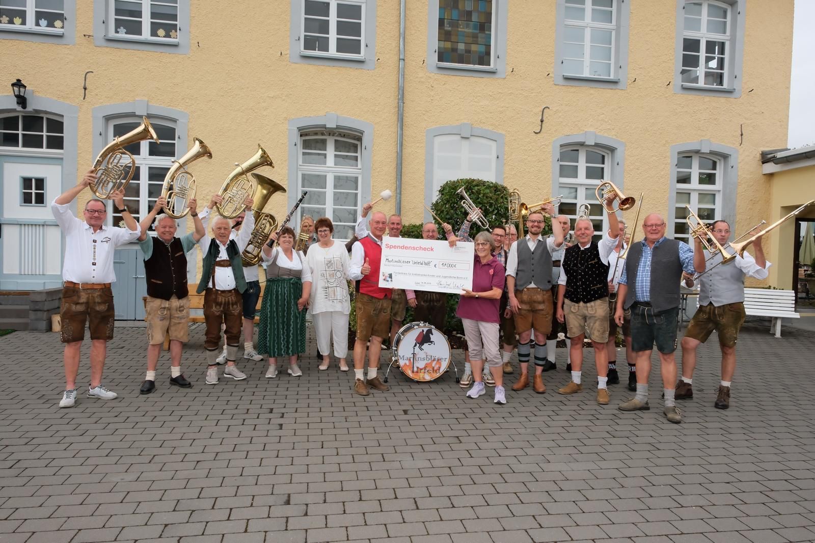Ein Mann und eine Frau halten einen großen Scheck, um sie herum stehen Männer und Frauen mit Musikinstrumenten.