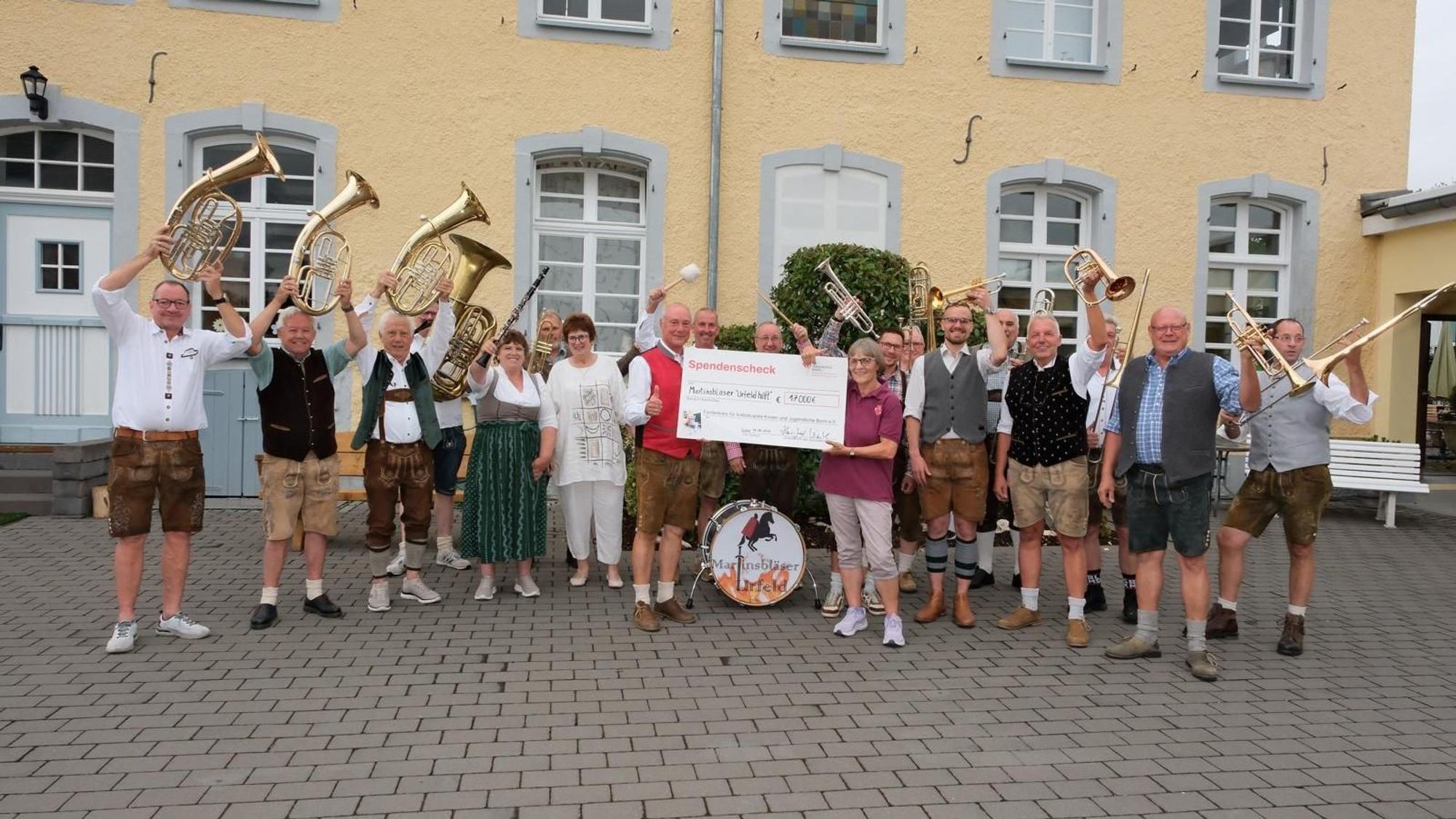 Ein Mann und eine Frau halten einen großen Scheck, um sie herum stehen Männer und Frauen mit Musikinstrumenten.