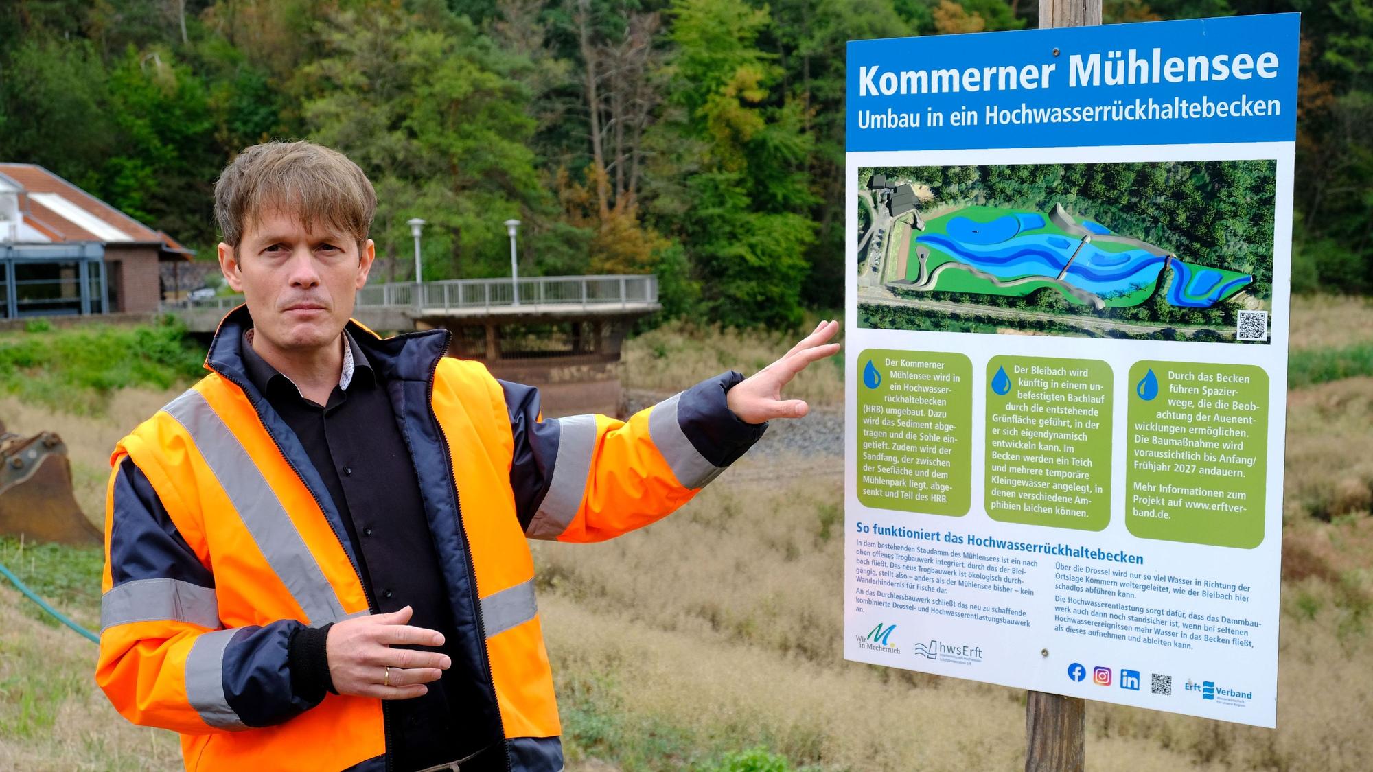 Ein Mann, der eine orangefarbene Signaljacke trägt, deutet auf eine Schautafel mit der Aufschrift „Kommerner Mühlensee - Umbau in ein Hochwasserrückhaltebecken“.