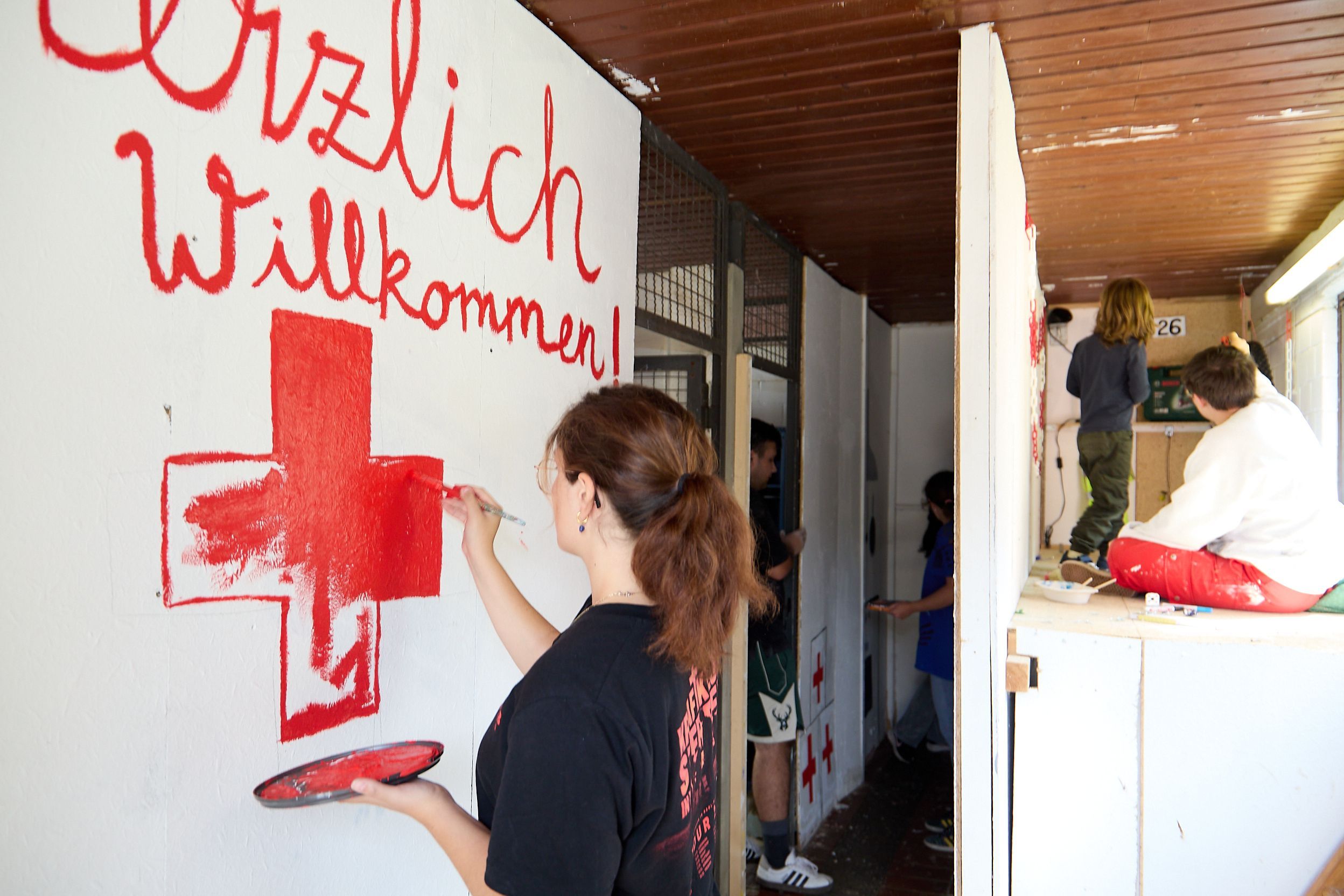 Eine junge Frau pinselt mit roter Frabe ein Kreuz auf eine weiße Wand. Darüber ist der Schriftzug „Herzlich Willkommen“ zu sehen.