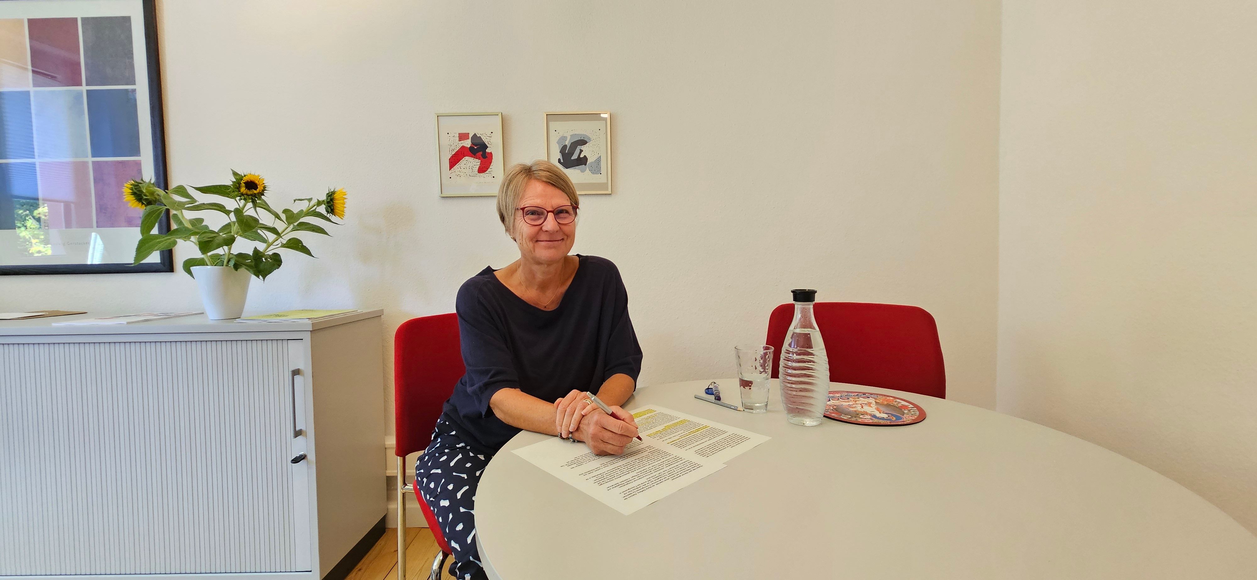 Auf dem Bild ist das Portrait einer Frau zu sehen, sie sitzt an einem Tisch.