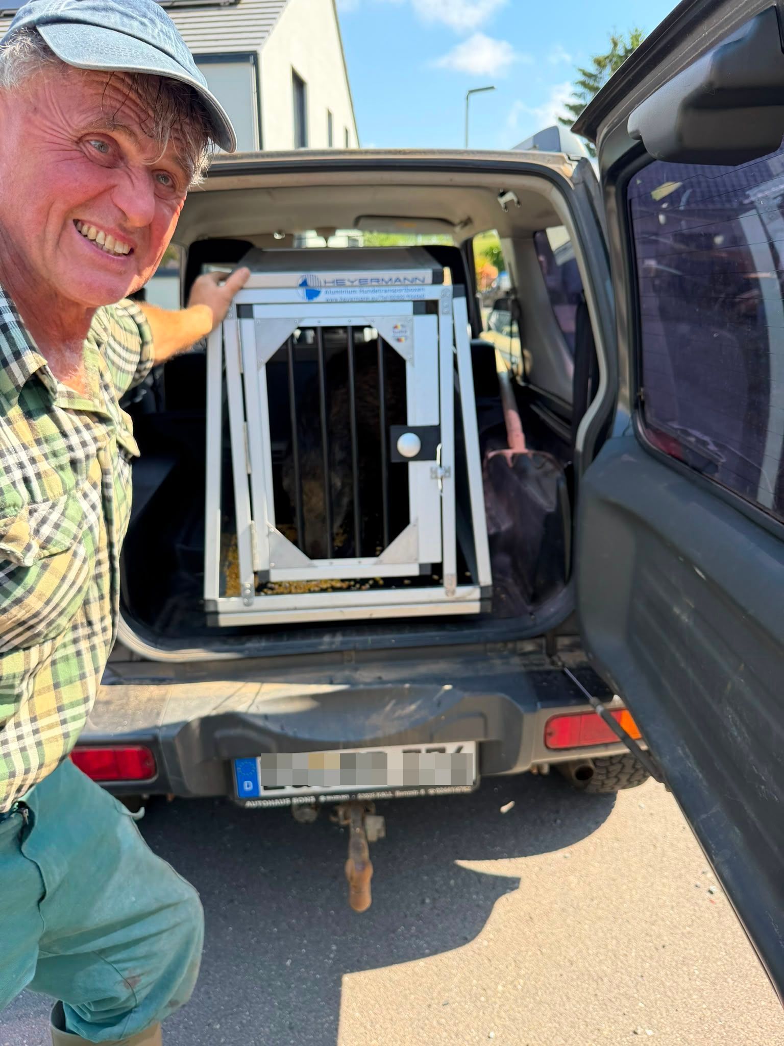 Ein Mann im karierten Hemd steht neben der geöffneten Hecktür eines Jeeps, in dem sich eine Hundetransportbox befindet.