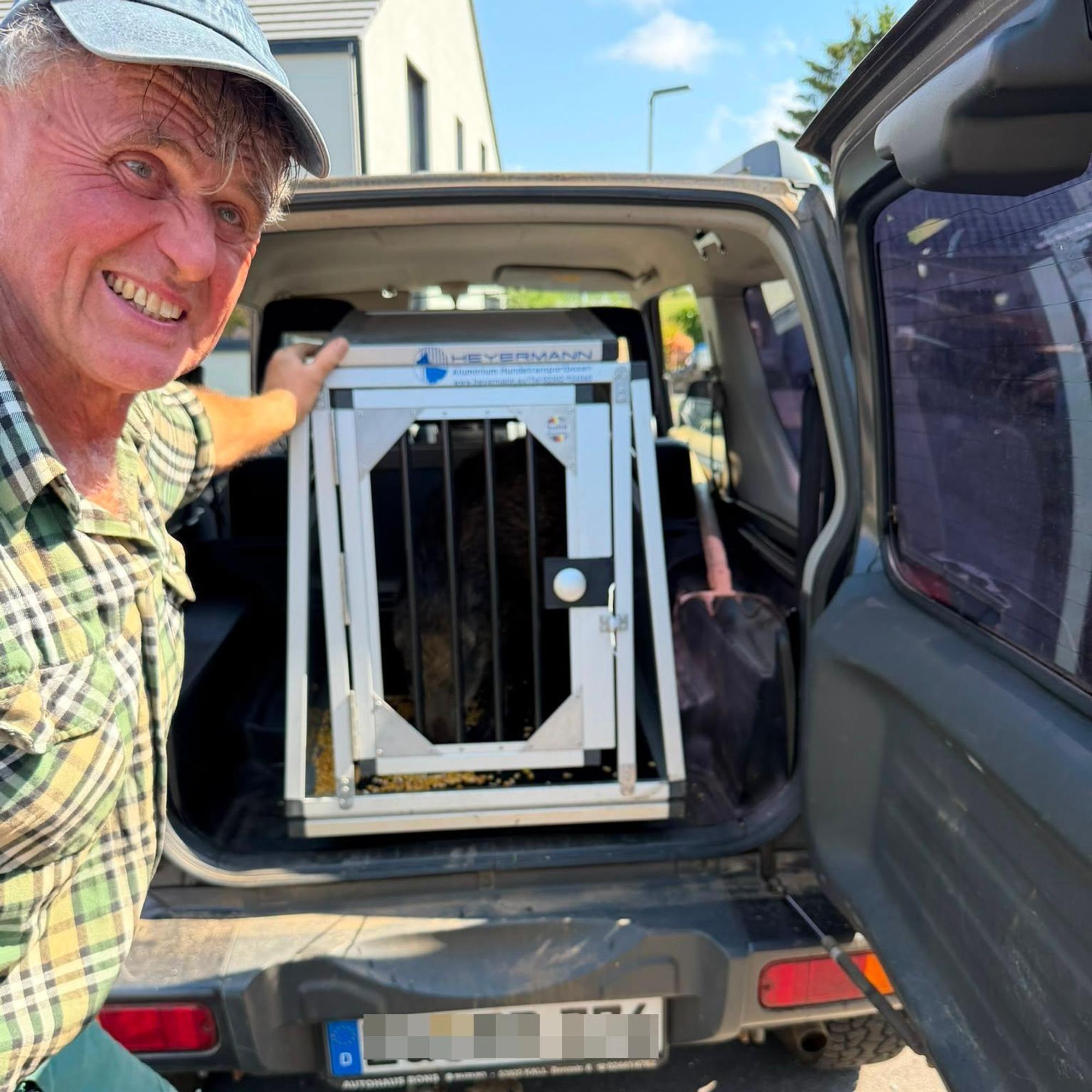 Ein Mann im karierten Hemd steht neben der geöffneten Hecktür eines Jeeps, in dem sich eine Hundetransportbox befindet.