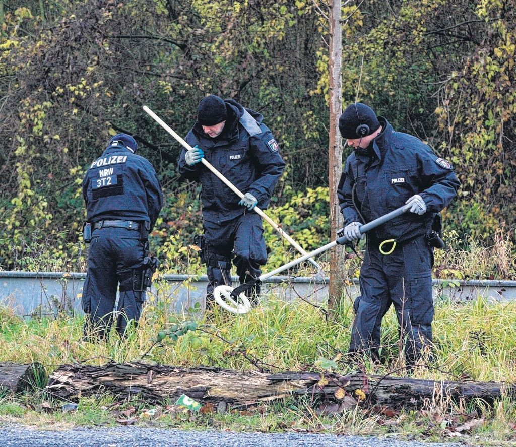 Zu sehen sind drei Polizisten, einer hält einen Metalldetektor, ein anderer eine Harke.