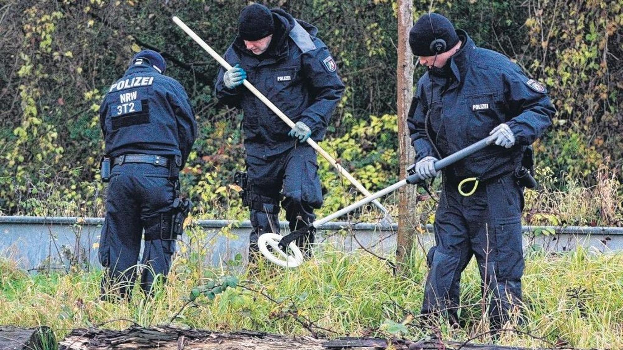 Zu sehen sind drei Polizisten, einer hält einen Metalldetektor, ein anderer eine Harke.