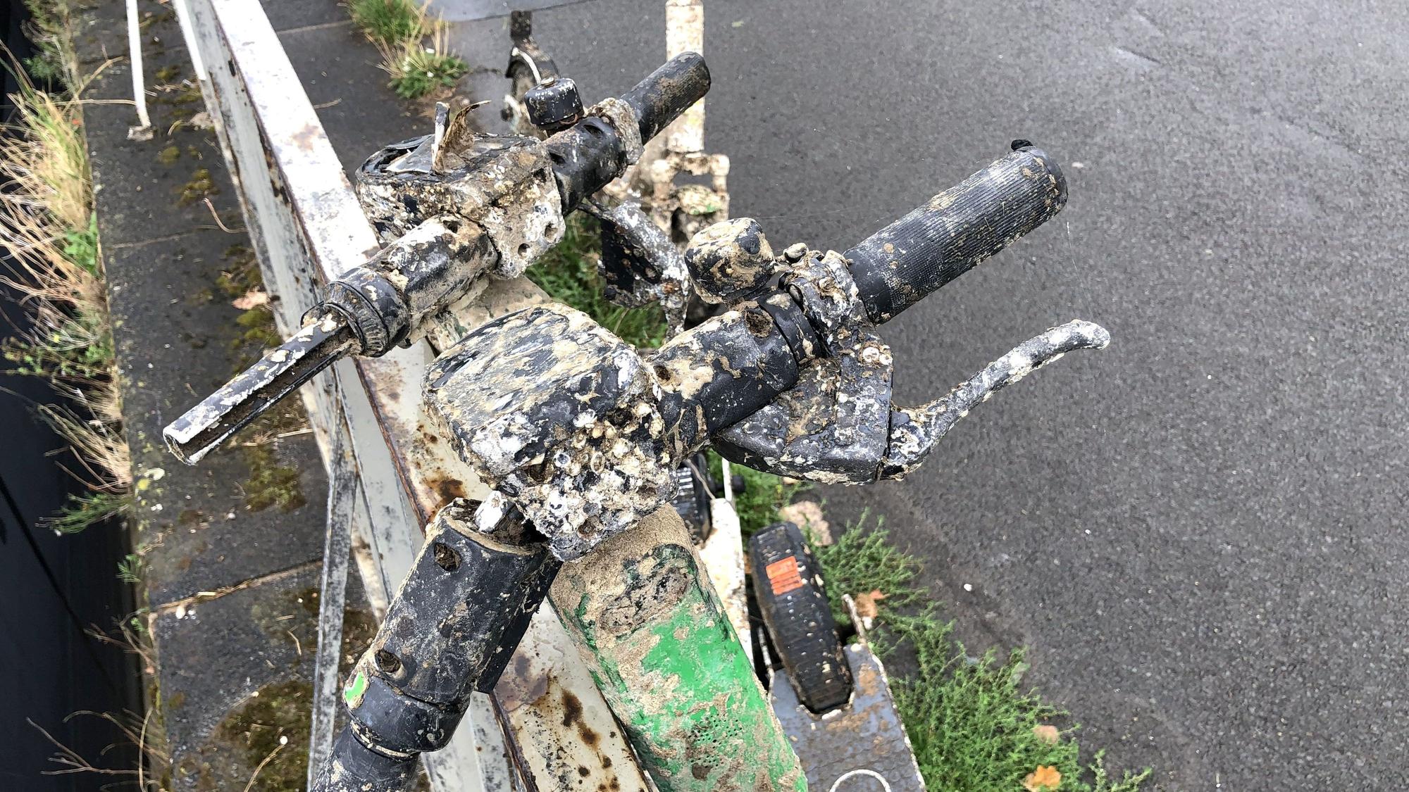 Defekte und mit getrocknetem Schlamm überzogene E-Scooter am Rheinufer in Köln-Mülheim.