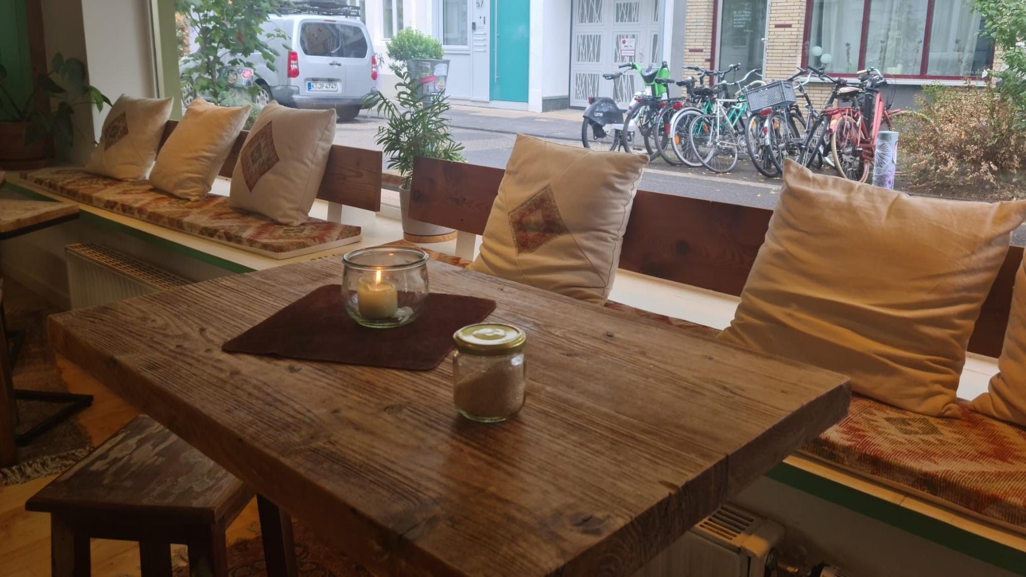 Schlichte Holztische und Sitzbänke stehen vor einer langen Fensterfront, durch die man auf eine Straße sieht, an der viele Fahrräder parken.