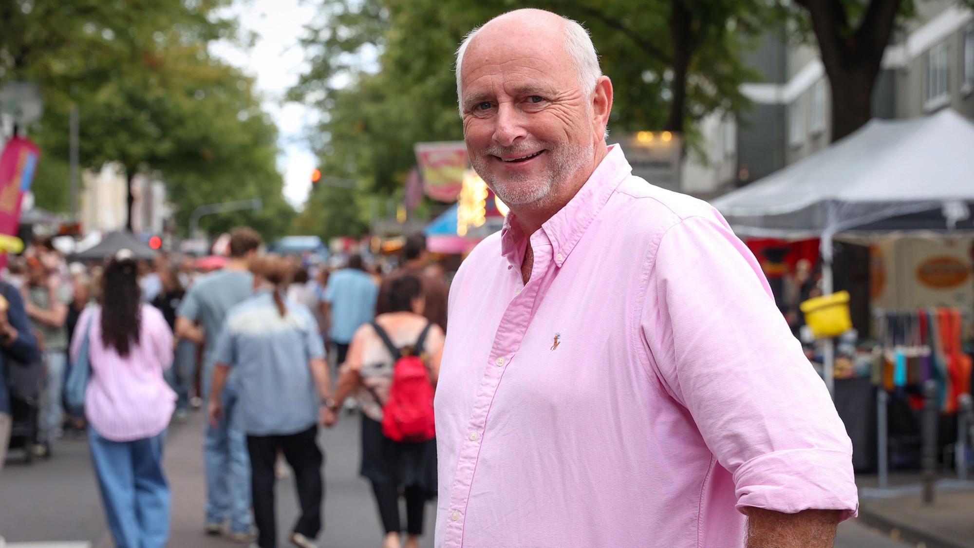 Ein Mann mit Bart und rosa Shirt steht auf einer Straße, auf der Stände aufgebaut sind und viele Menschen unterwegs sind.