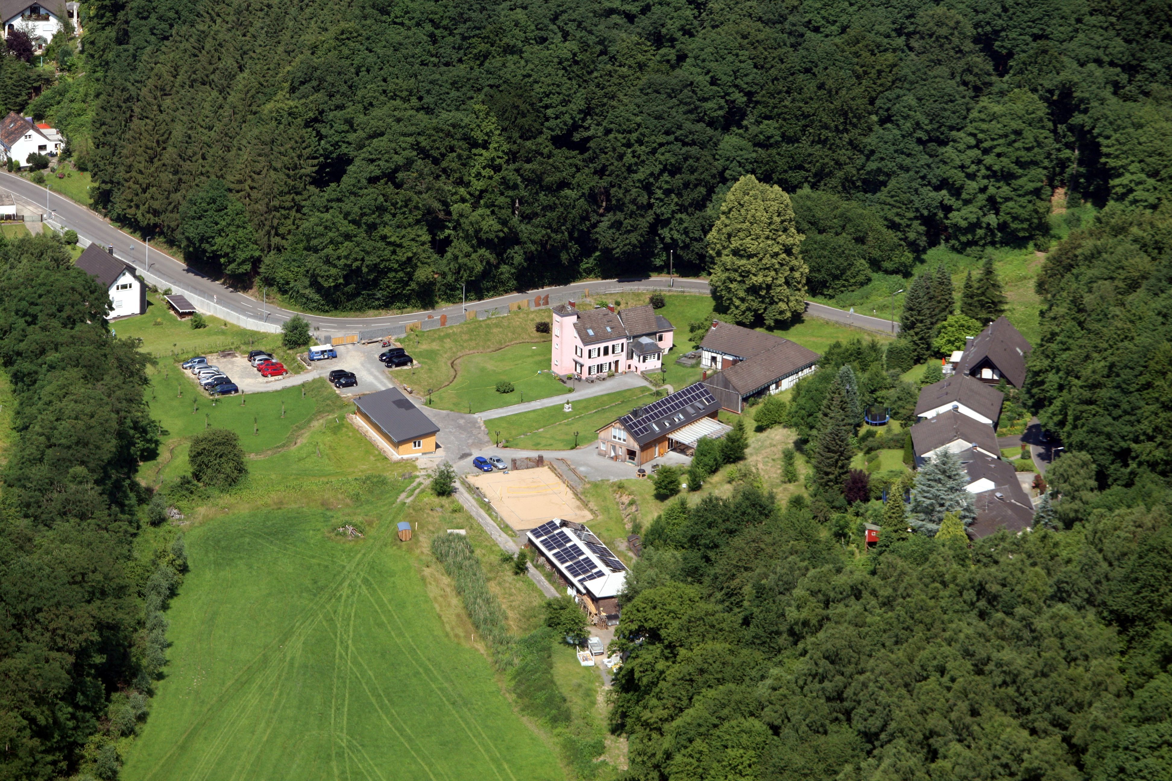 Eine Luftaufnahme mit viel Wiese und Bäumen, einer Straße und wenigen Gebäuden