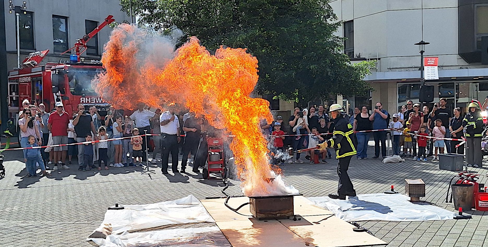 Ein Feuerwehrmann löscht eine hohe Stichflamme, Menschen hinter einer Absperrung schauen zu.