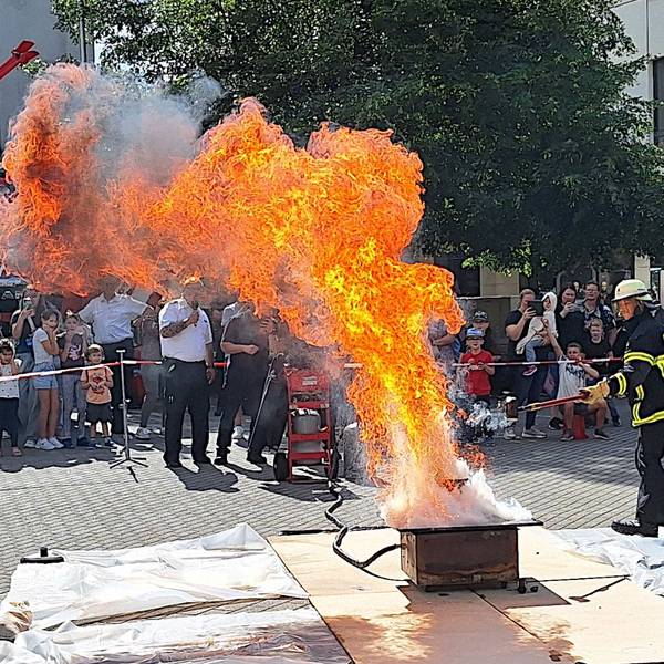 Ein Feuerwehrmann löscht eine hohe Stichflamme, Menschen hinter einer Absperrung schauen zu.