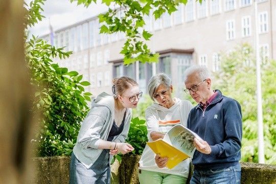 ZWei Seniorenstudenten und eie junge Frau schauen in eine Broschüre, im Hintergrund sieht man das Hauptgebäude der Uni Köln.