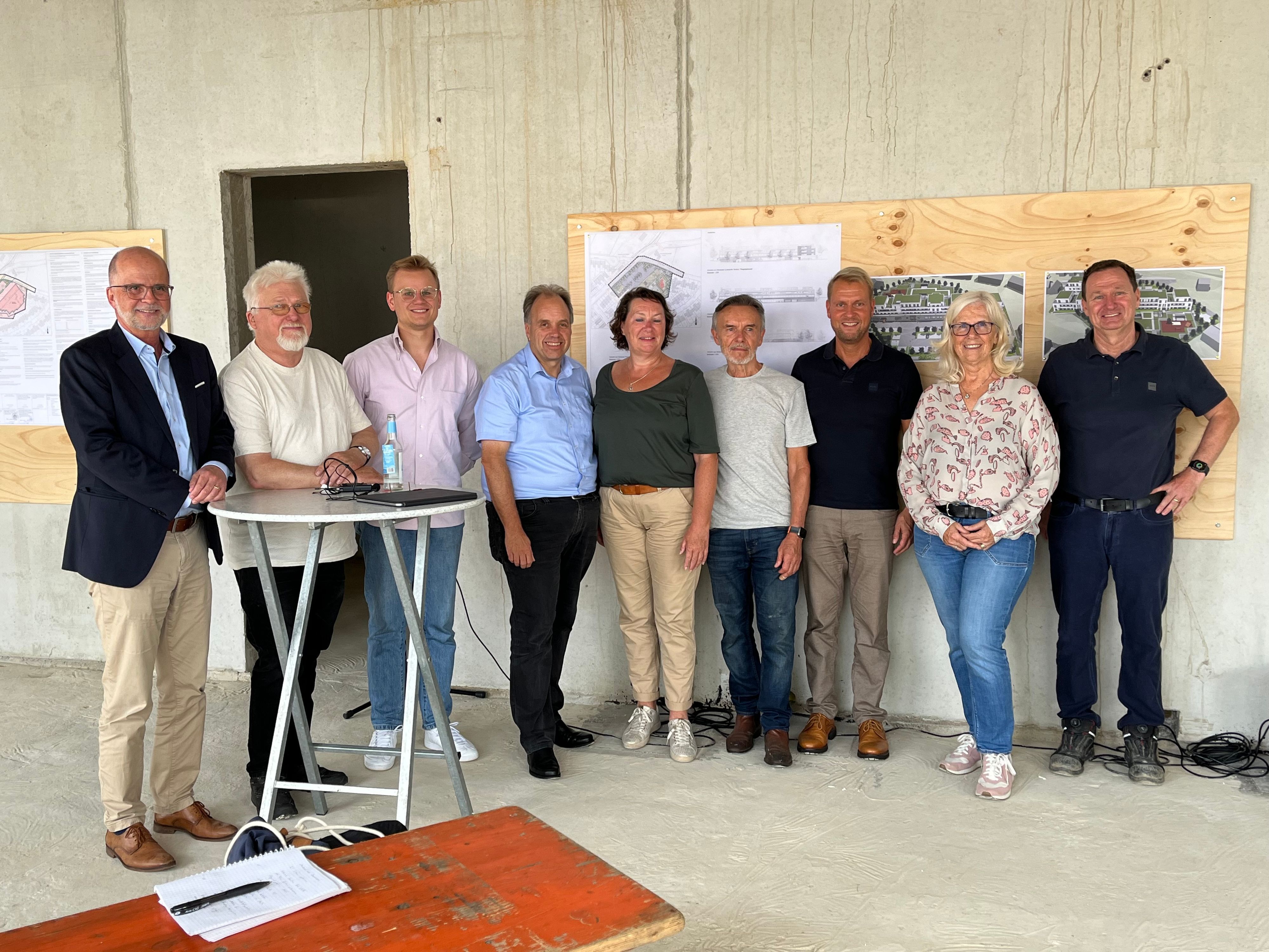 Männer und Frauen stehen nebeneinander an einer Wand, an der Baupläne befestigt sind. Zwei Männer stehen an einem Stehtisch. Sie befinden sich in einem Rohbau.