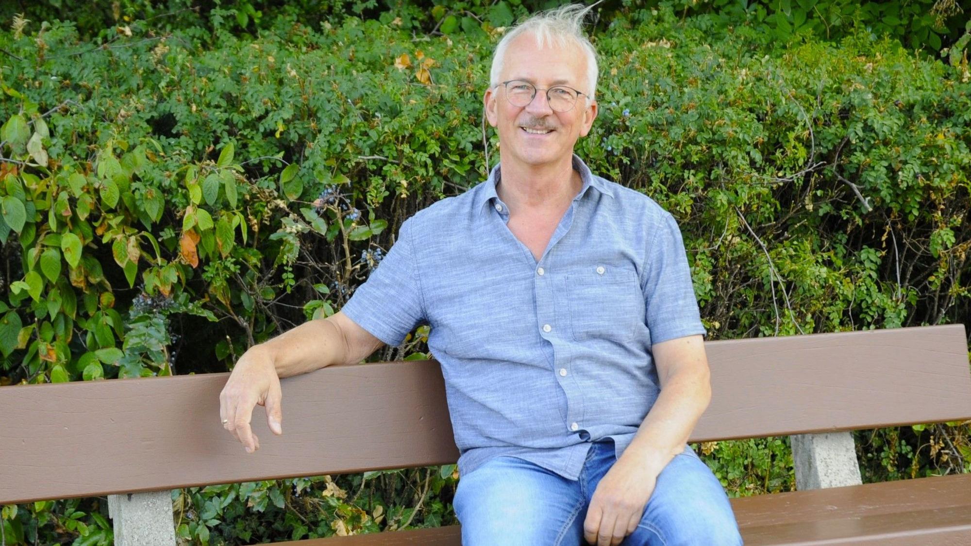 Das Bild zeigt einen Mann mit Brille und grauen Haaren, der auf einer Bank sitzt.