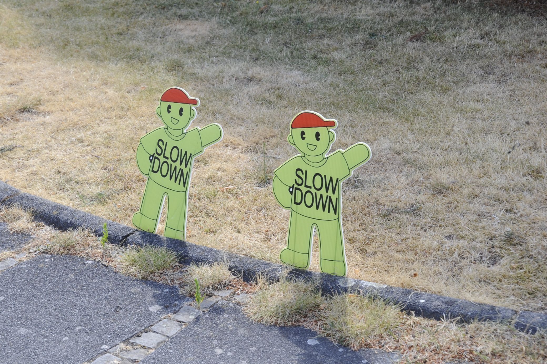 Das Bild zeigt kleine grüne Figuren mit roten Kappen an einer Straße, auf denen „Slow down“ steht.