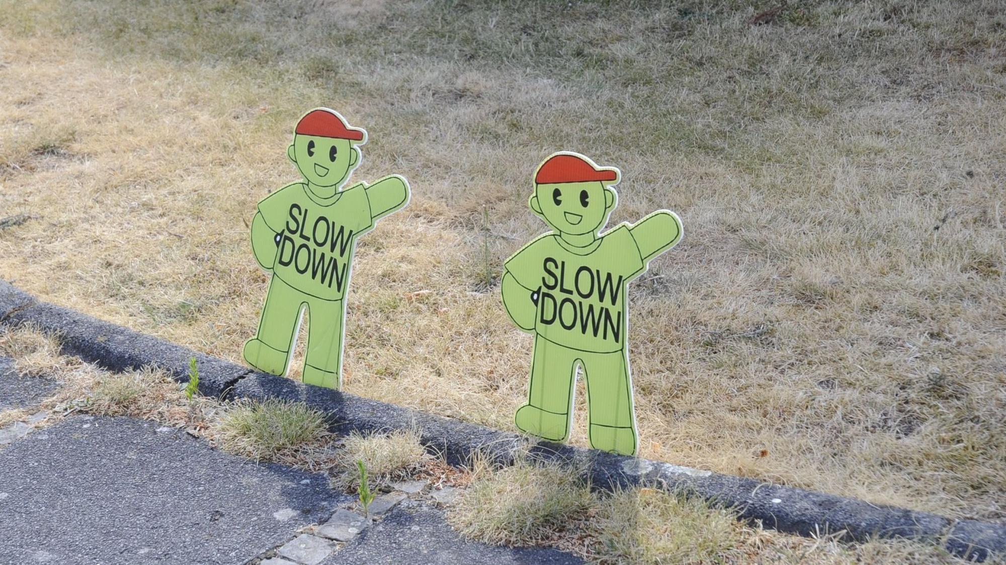 Das Bild zeigt kleine grüne Figuren mit roten Kappen an einer Straße, auf denen „Slow down“ steht.